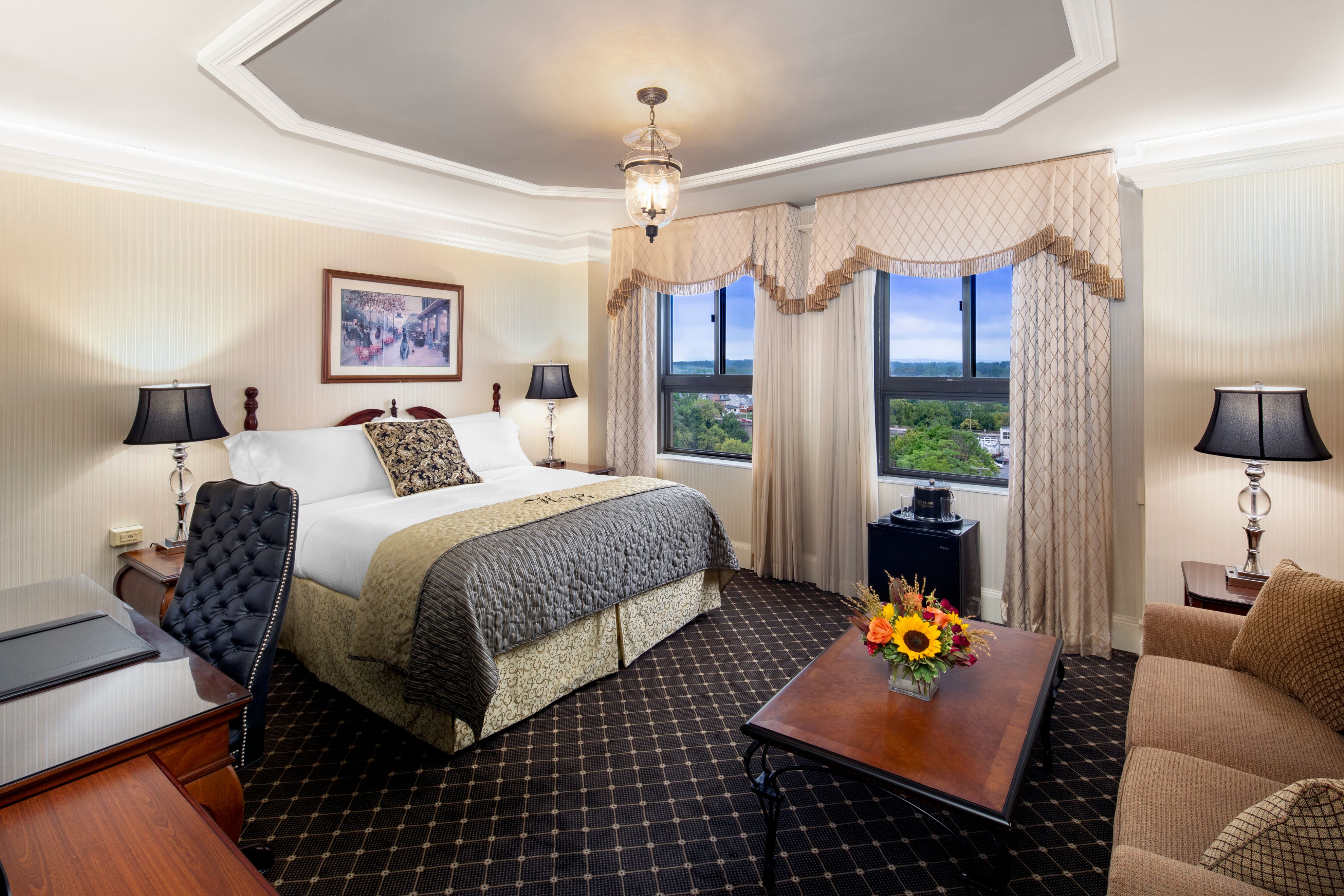 An elegant hotel bedroom featuring a king-size bed with a gold coverlet, a dark wood coffee table with flowers, a beige sofa, and windows with fringed drapes looking out over trees.