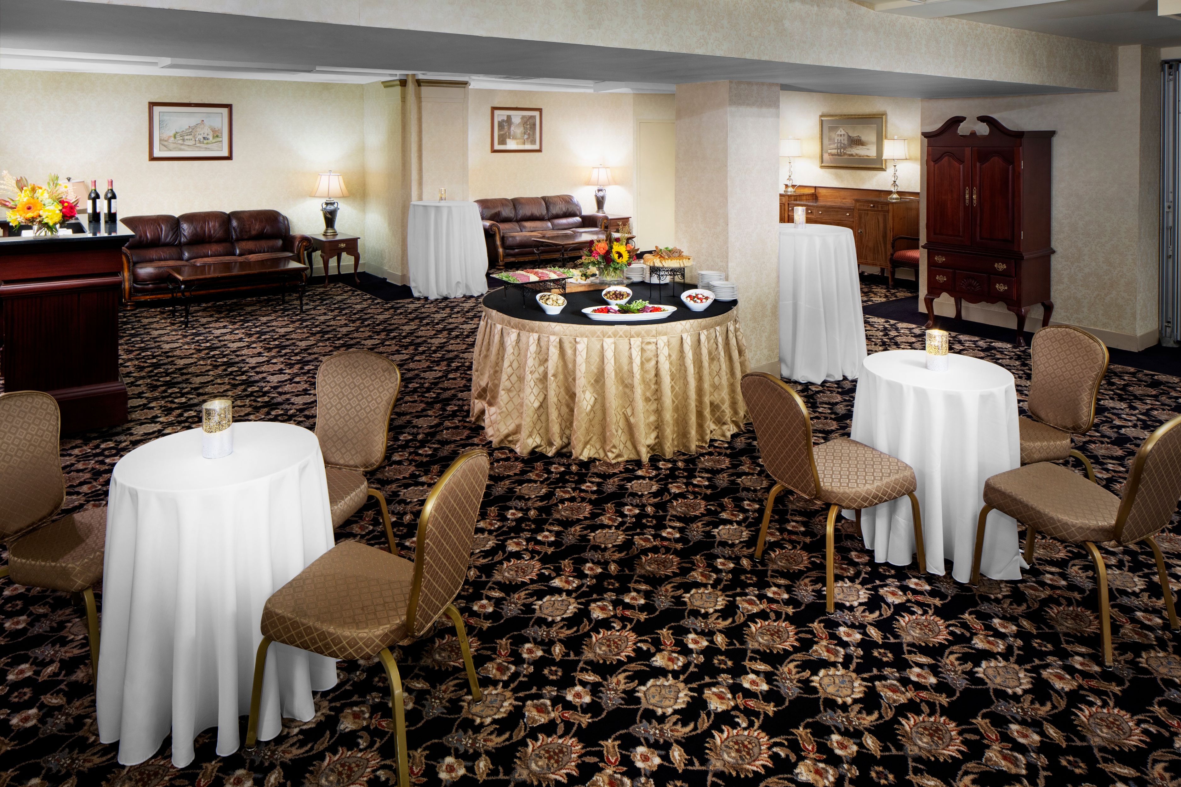 A banquet hall lounge set up for an event with round tables covered in white linens, a central buffet table, brown leather couches, and patterned carpet.