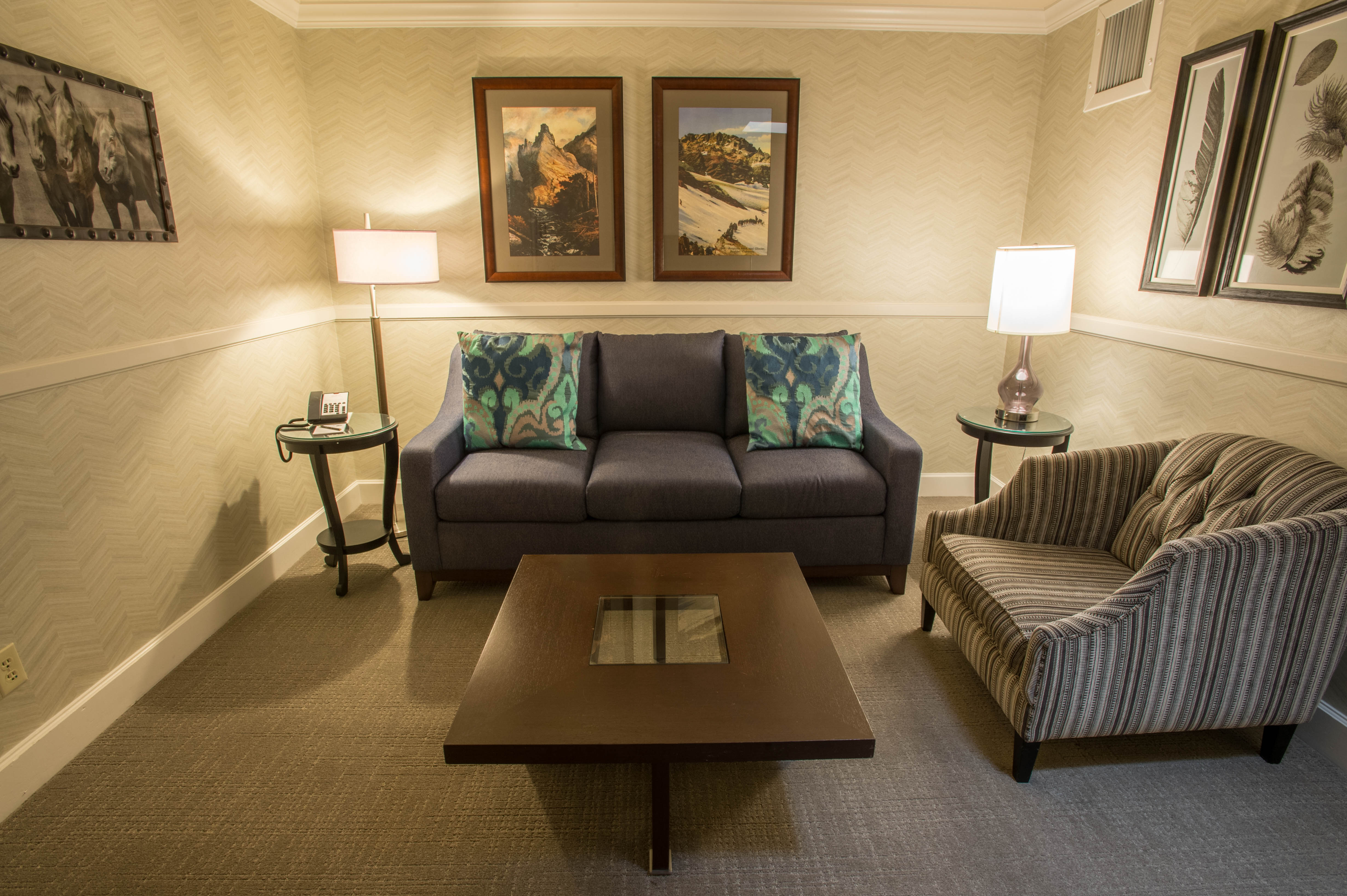 Cozy hotel sitting area featuring a dark blue sofa with teal accent pillows, a striped armchair, a square wooden coffee table, and framed artwork on light patterned walls.