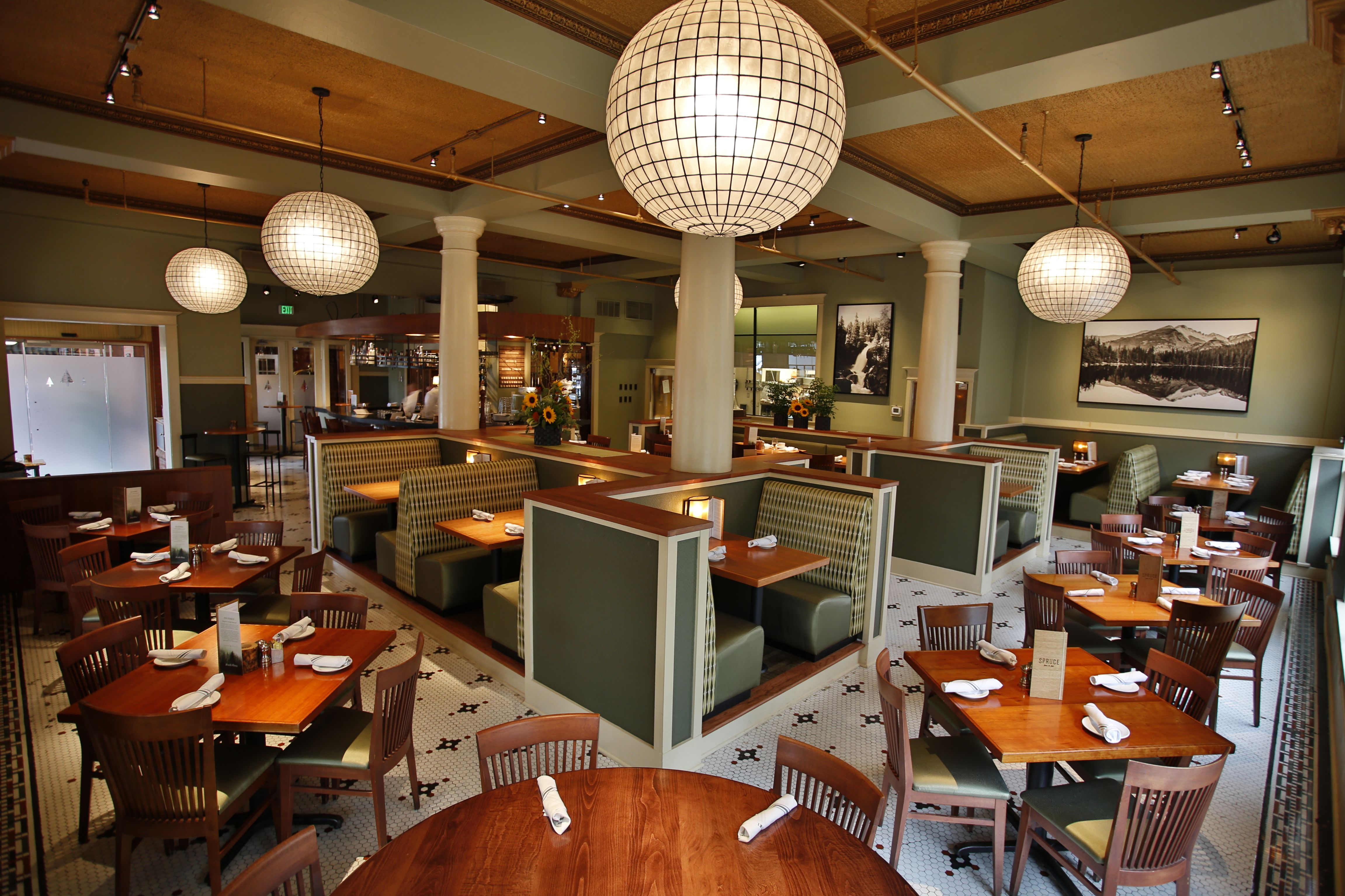 Spacious hotel restaurant featuring green booth seating, warm wooden tables and chairs, large spherical pendant lights, tiled flooring, and nature landscape artwork on olive-green walls.