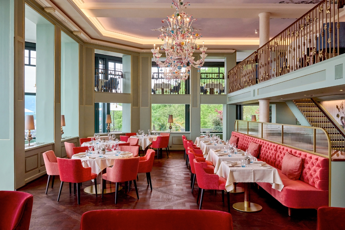 Spacious, high-ceilinged hotel restaurant with a mezzanine level, featuring light green walls, a colorful floral glass chandelier, red dining chairs, and a long tufted red velvet banquette along wood parquet flooring.