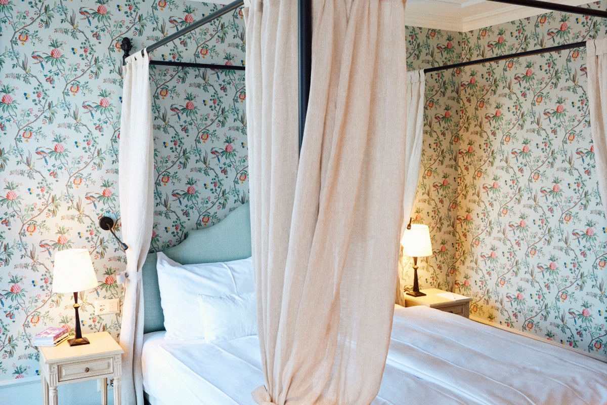Cozy, vintage-inspired hotel bedroom with floral botanical wallpaper, featuring a black metal four-poster canopy bed draped with linen curtains and flanked by bedside tables with glowing lamps.