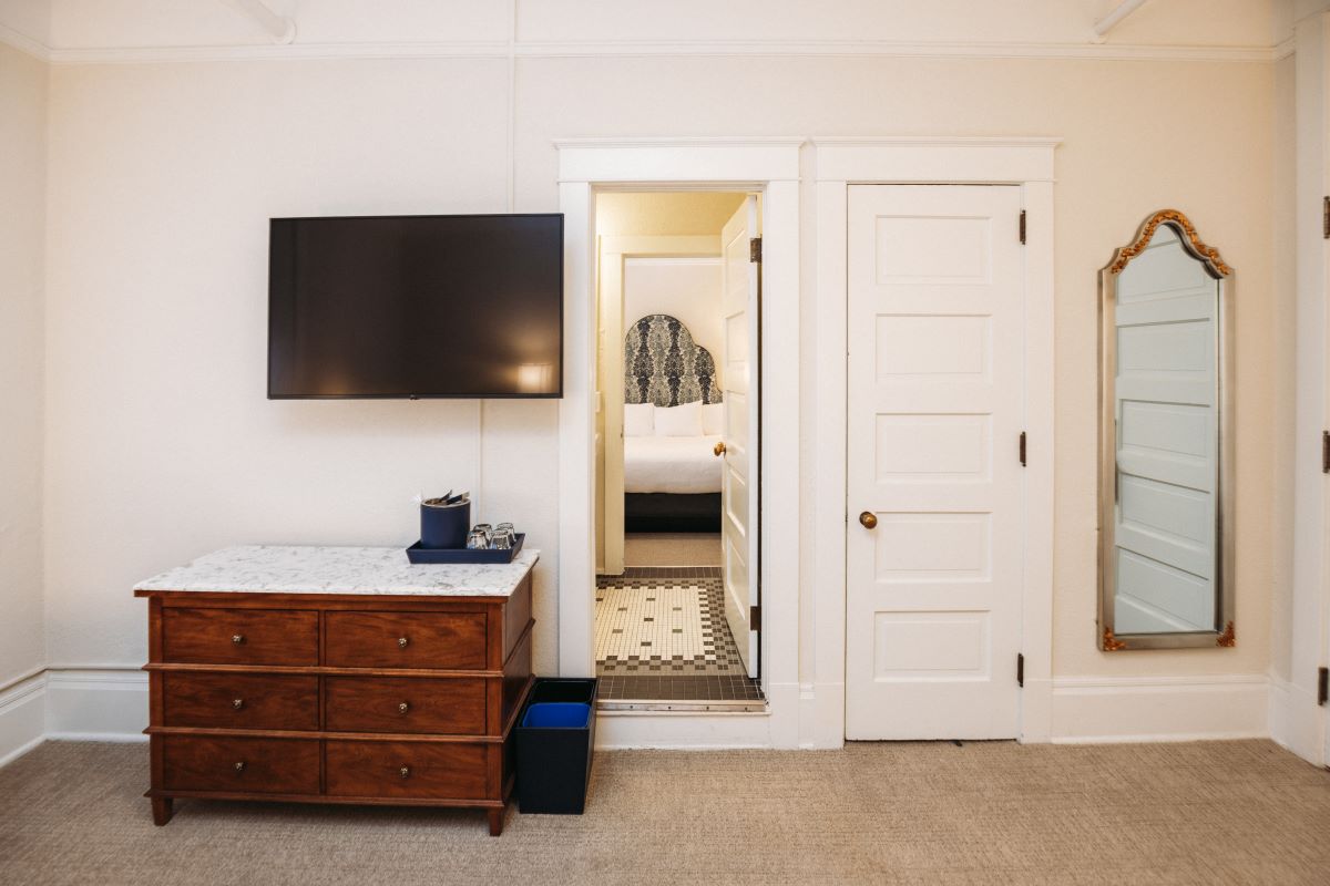 Hotel room interior with neutral walls, a marble-topped wooden dresser under a wall-mounted TV, a doorway leading into a bathroom with tile flooring, an adjacent bedroom, and a full-length wall mirror with a ornate gold trim.