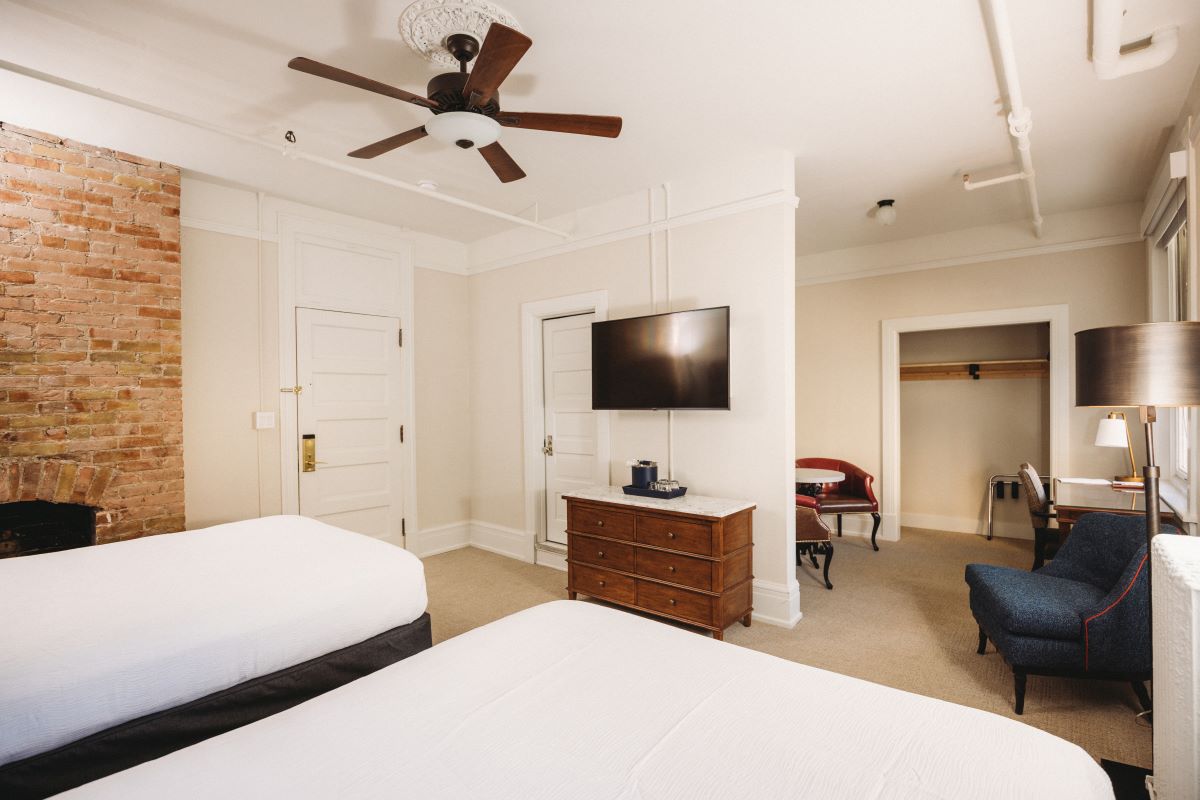 Spacious hotel room featuring two beds with white linens, an exposed brick wall, a ceiling fan, a wall-mounted TV over a wooden dresser, and a small sitting area with an armchair and desk.