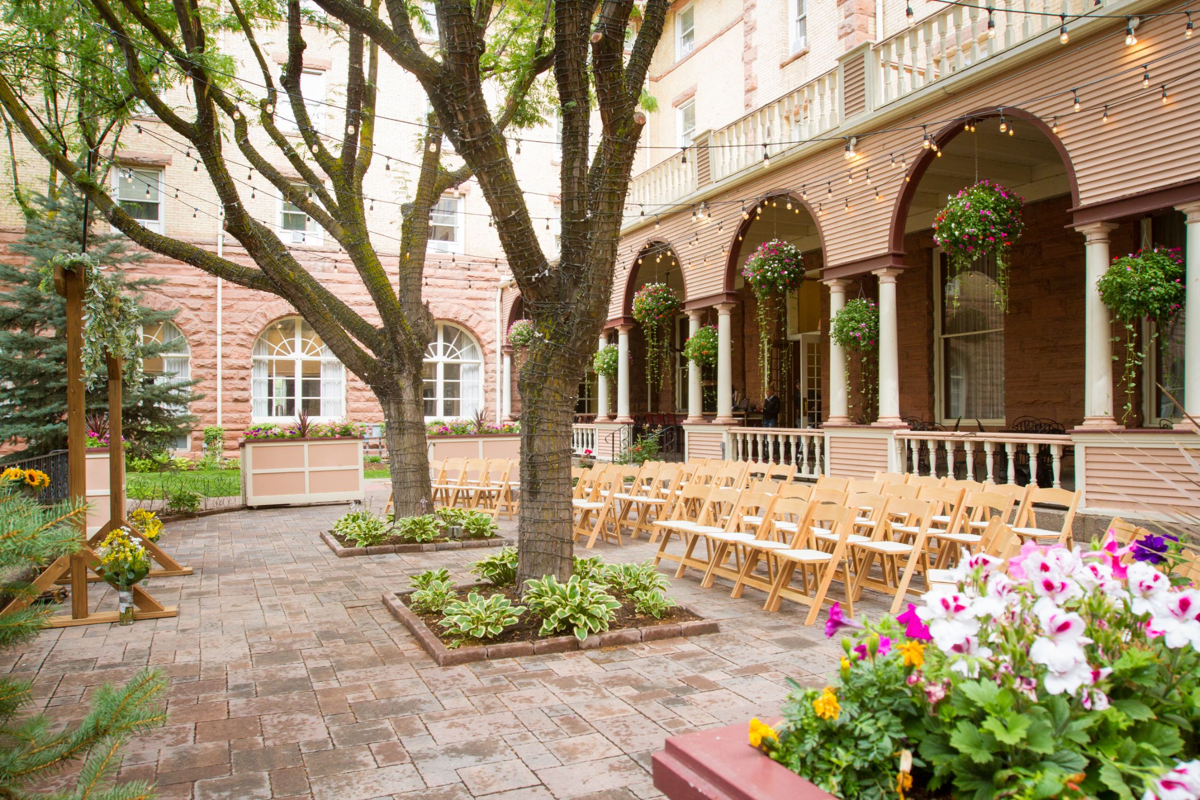 An outdoor paved courtyard set up for a wedding ceremony, featuring rows of light wooden folding chairs, mature trees wrapped in string lights, hanging flower baskets along a covered colonnade, and vibrant potted flowers in the foreground.