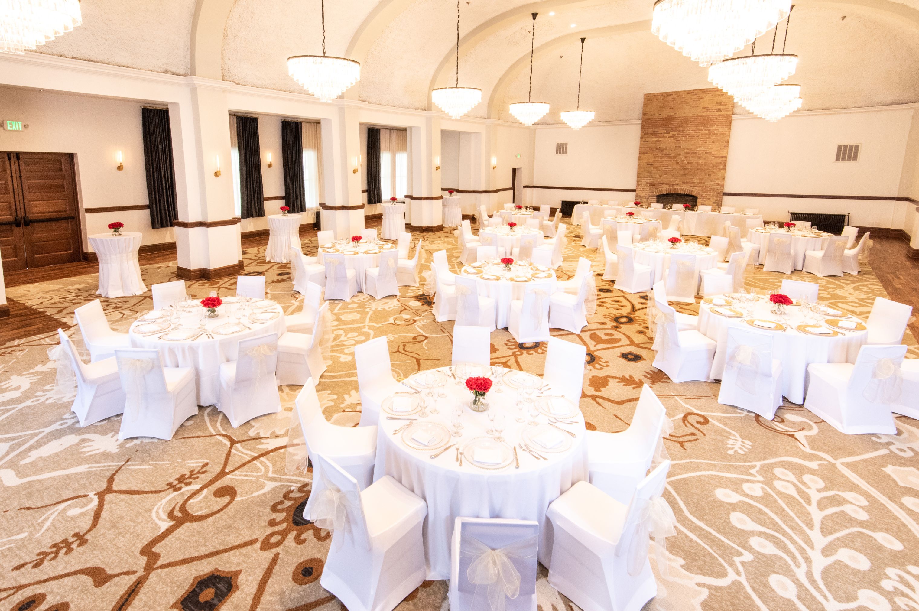 Grand event hall set for a banquet with round tables, white table linens, white chair covers, red floral centerpieces, crystal chandeliers, and a large brick fireplace in the background.