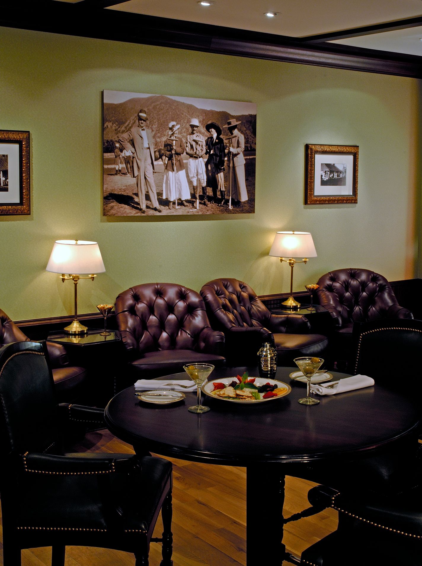 Sophisticated upscale lounge featuring dark leather tufted armchairs lined against light sage green walls, a dark round dining table set with martinis and an appetizer platter, illuminated warm table lamps, and a large black-and-white vintage photograph o