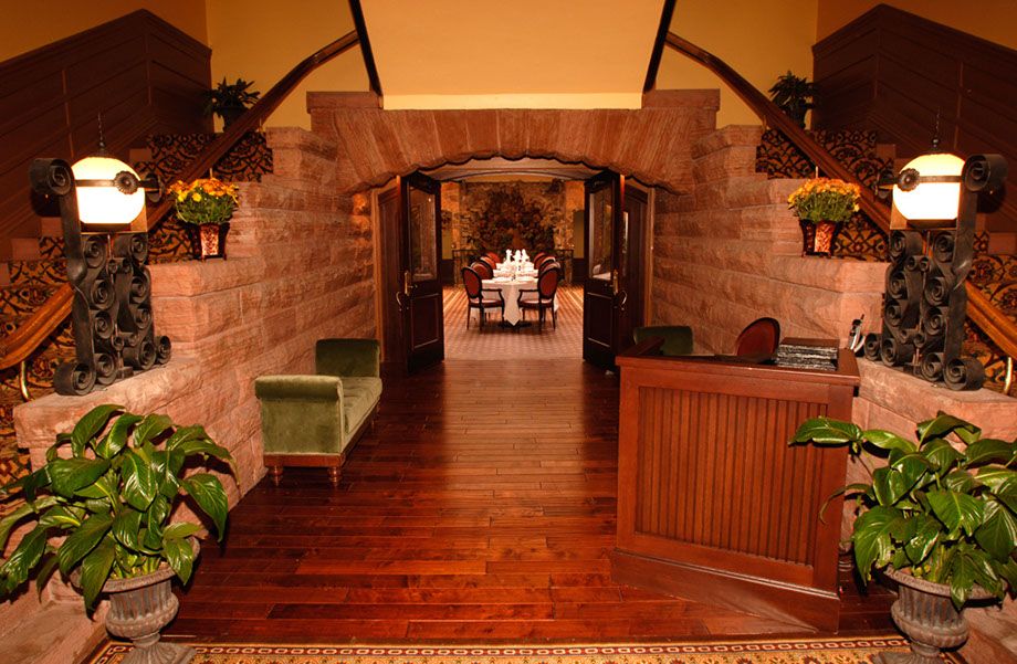 Grand entrance to an elegant dining room framed by red sandstone brick archways and dual staircases, featuring dark hardwood flooring, glowing orb lamps on ornate wrought iron stands, green potted plants, and a wooden host podium.