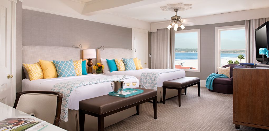 A bright hotel room with two double beds adorned with vibrant yellow and turquoise accent pillows, dark leather benches at the foot of each bed, and large windows looking out onto a sunny beach and ocean.