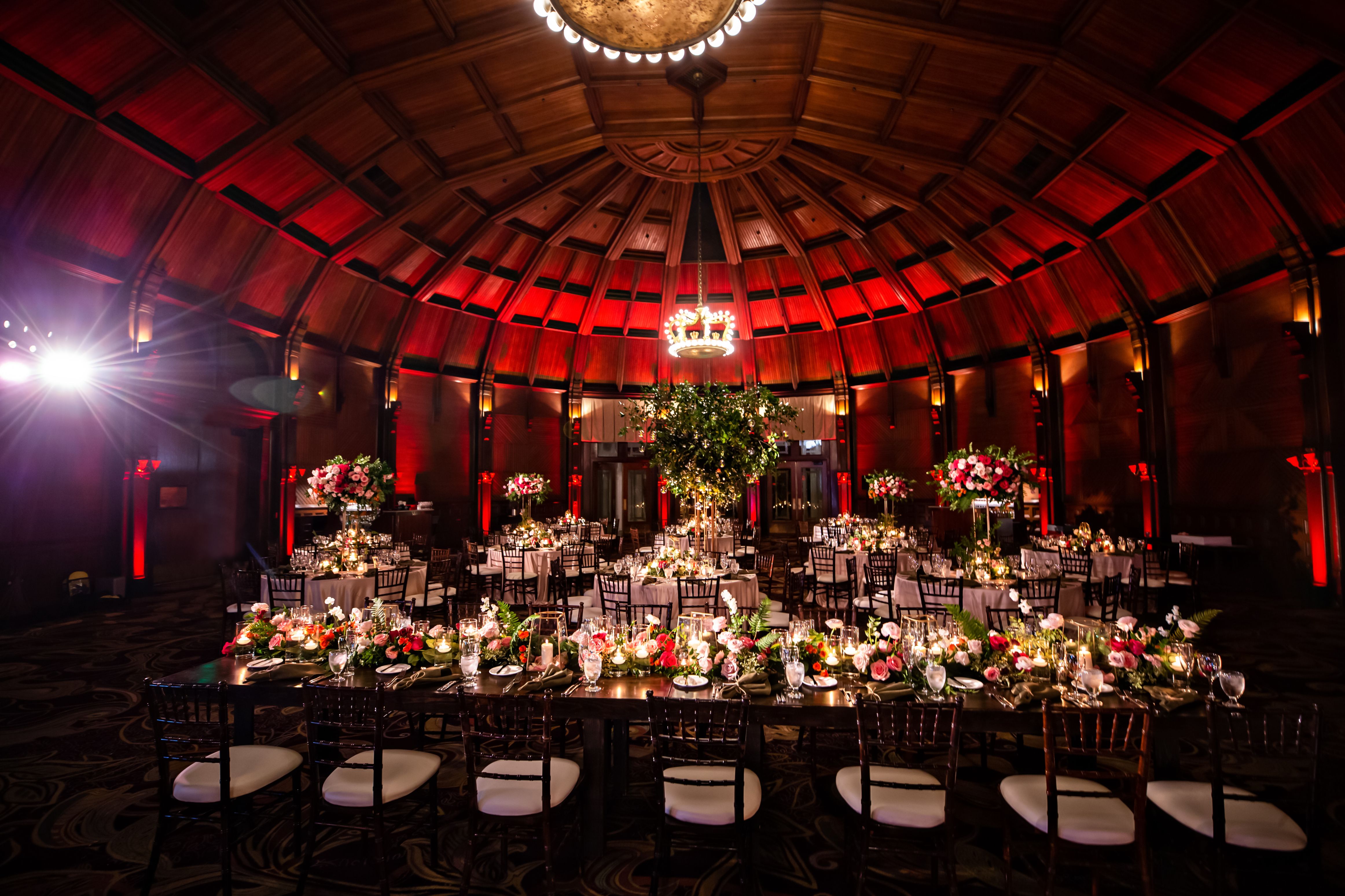 A lavish banquet room under a vaulted wooden dome ceiling lit with red ambient uplighting, featuring round and long wooden dining tables adorned with lush floral centerpieces, candles, and elegant table settings.