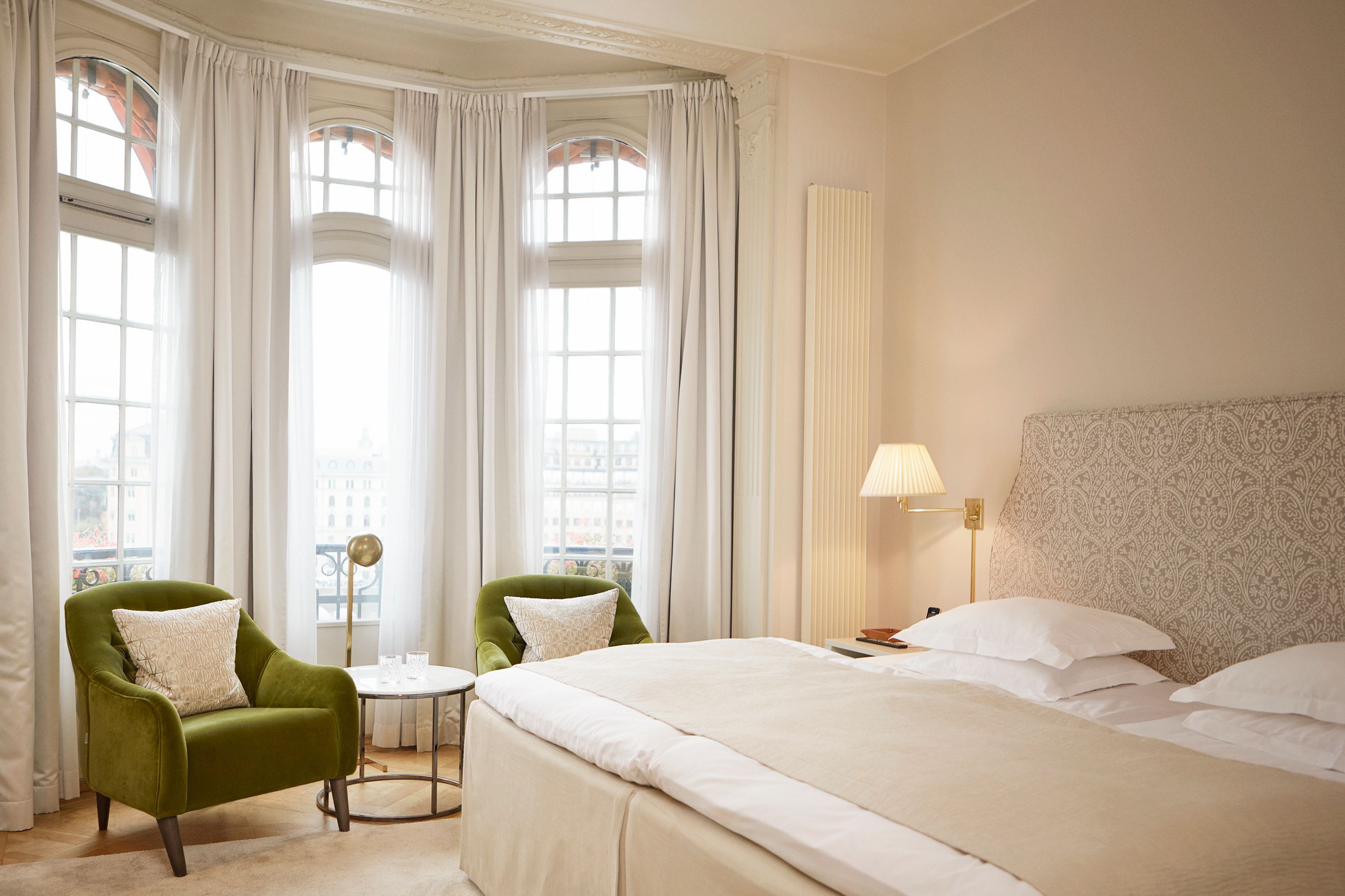 An elegant hotel bedroom featuring a bed with a patterned fabric headboard, two olive-green velvet armchairs positioned by a bay window with sheer white curtains, and soft neutral decor.