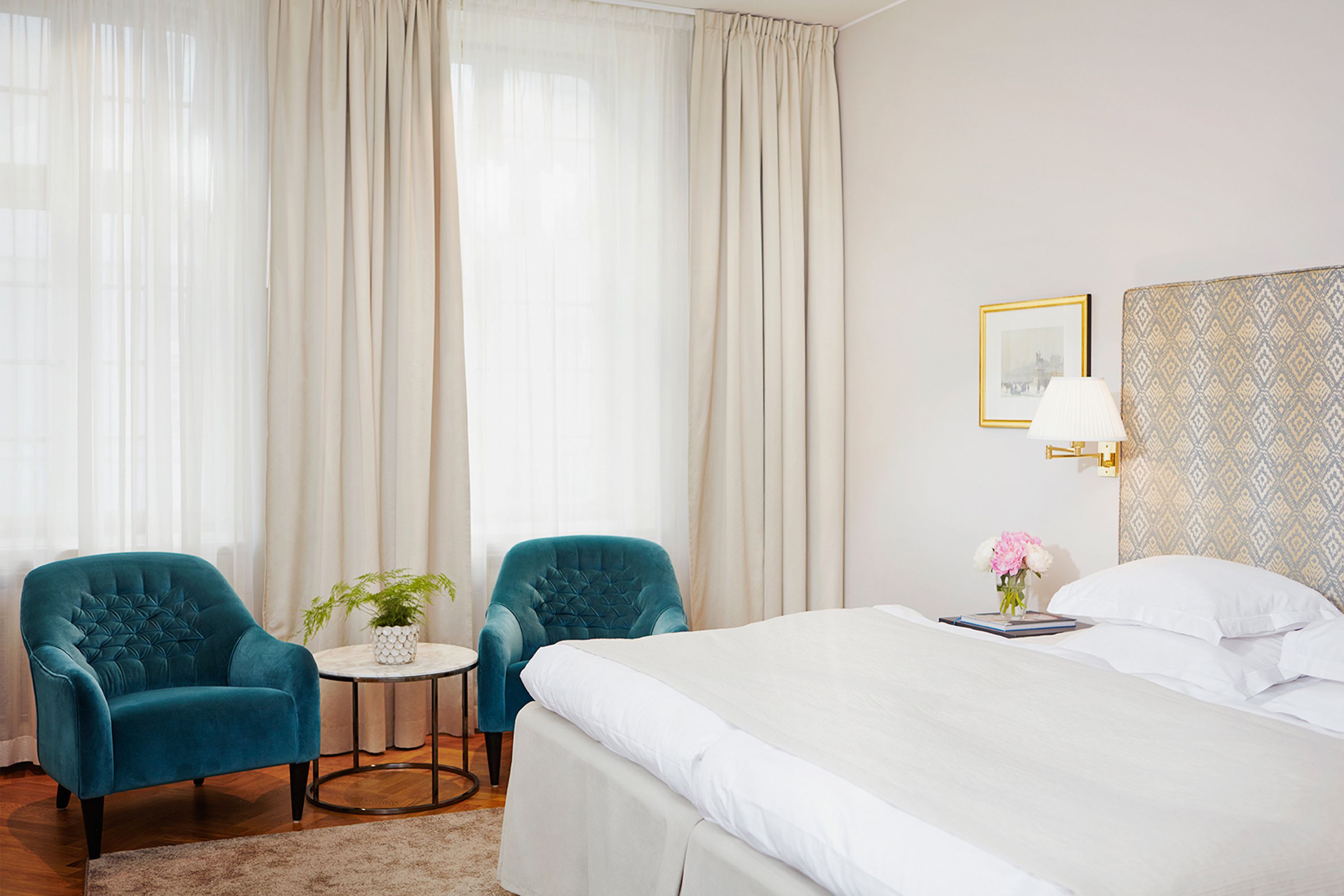 A brightly lit hotel room featuring a bed with a geometric-patterned headboard, white linens, and two plush teal velvet armchairs set beside a small round table near floor-to-ceiling cream curtains.