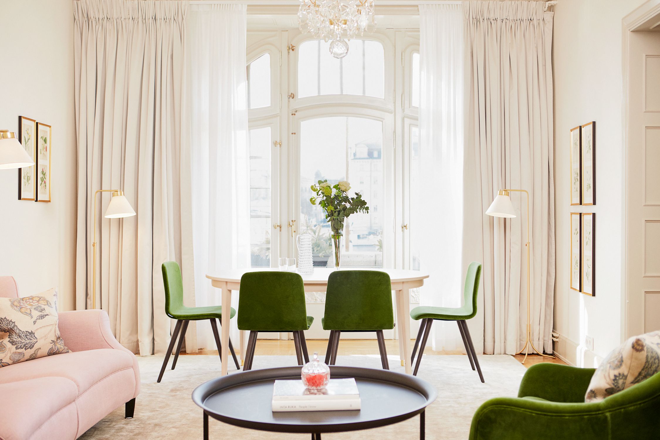 An elegant, sunlit living and dining room suite featuring green velvet chairs around a white dining table, framed by large arched windows with floor-to-ceiling white drapes, and soft pink sofa seating in the foreground.