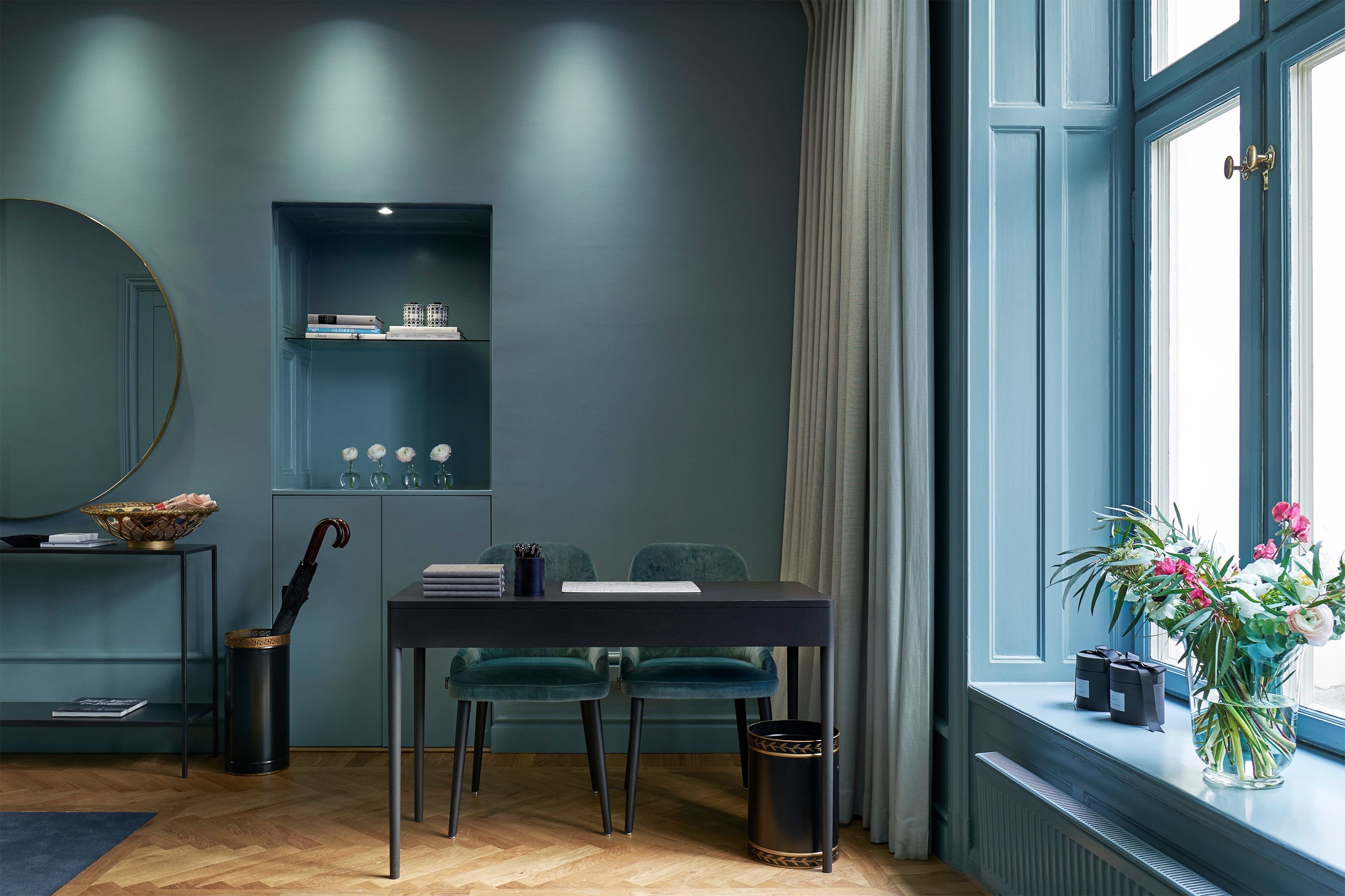 A sophisticated boutique office or reception room painted in moody teal-blue tones, featuring a black writing desk with two velvet chairs, a round mirror, and a window sill decorated with a vibrant flower arrangement.