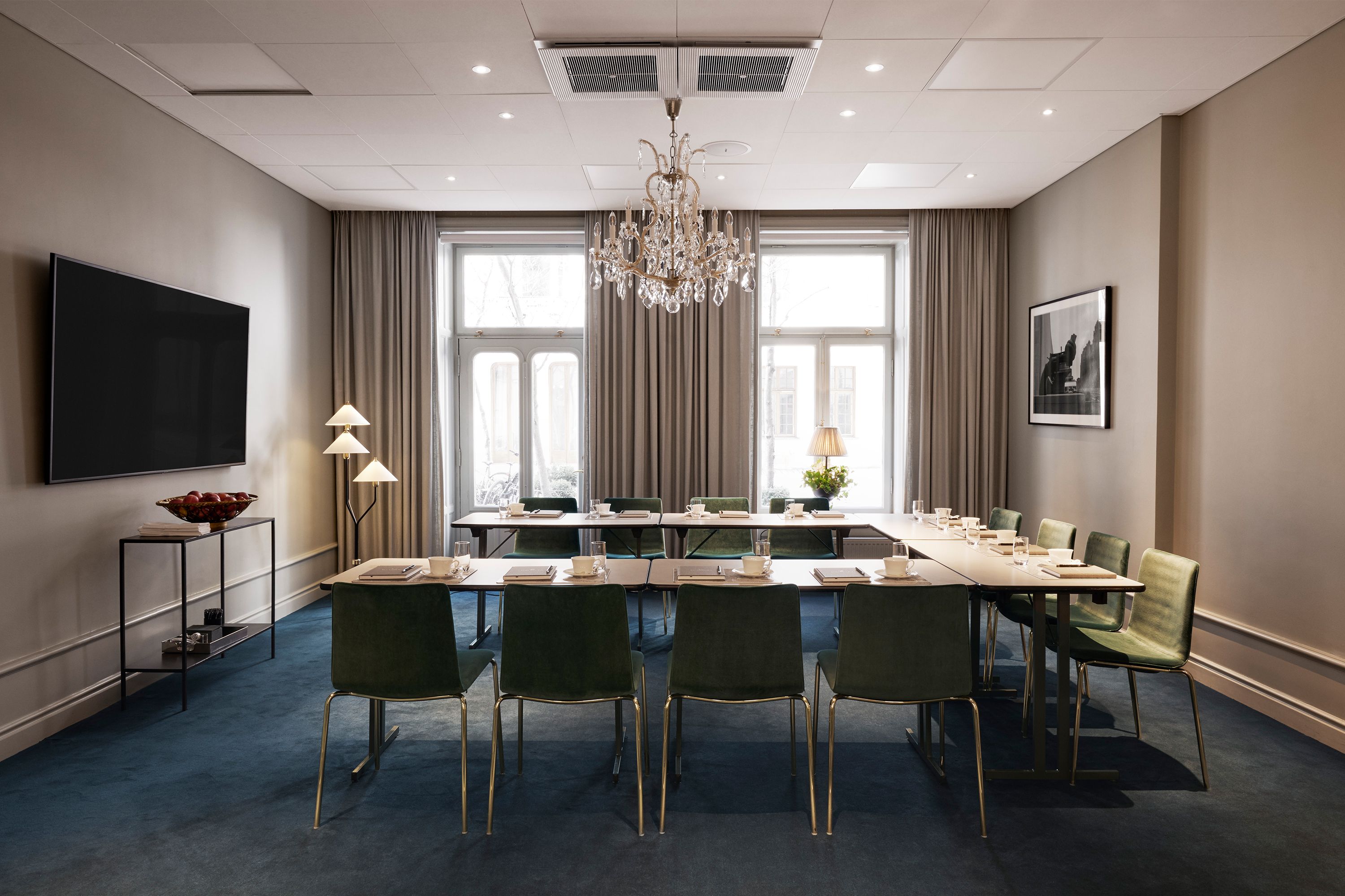 A modern, upscale hotel conference room arranged in a U-shape layout with green velvet dining chairs, a crystal chandelier hanging above, a wall-mounted TV, and long neutral drapes over the windows.