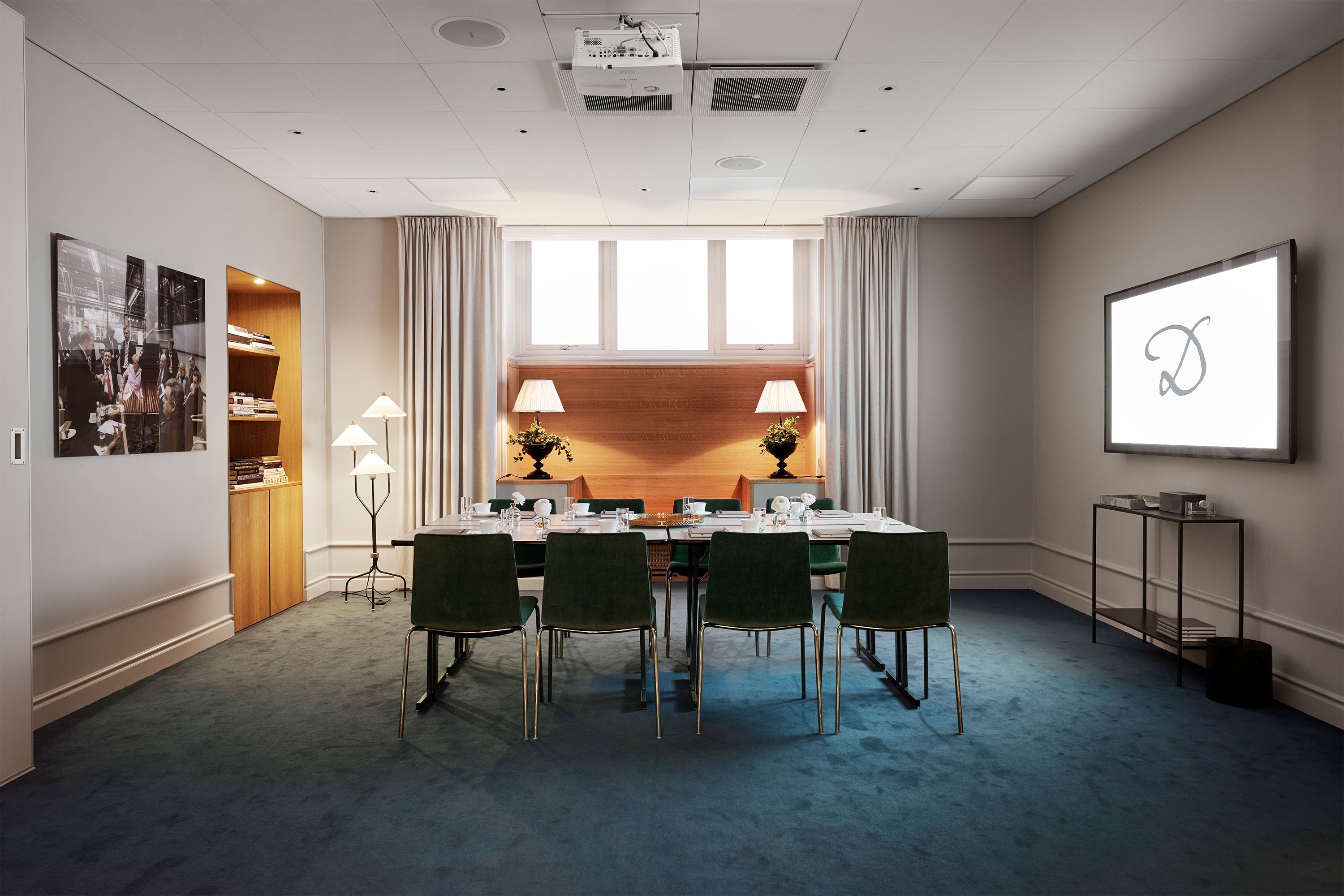 An intimate hotel boardroom set up for a meeting with green velvet chairs surrounding a long table, featuring warm ambient lighting, modern AV equipment, teal carpeting, and a recessed wooden bookshelf.