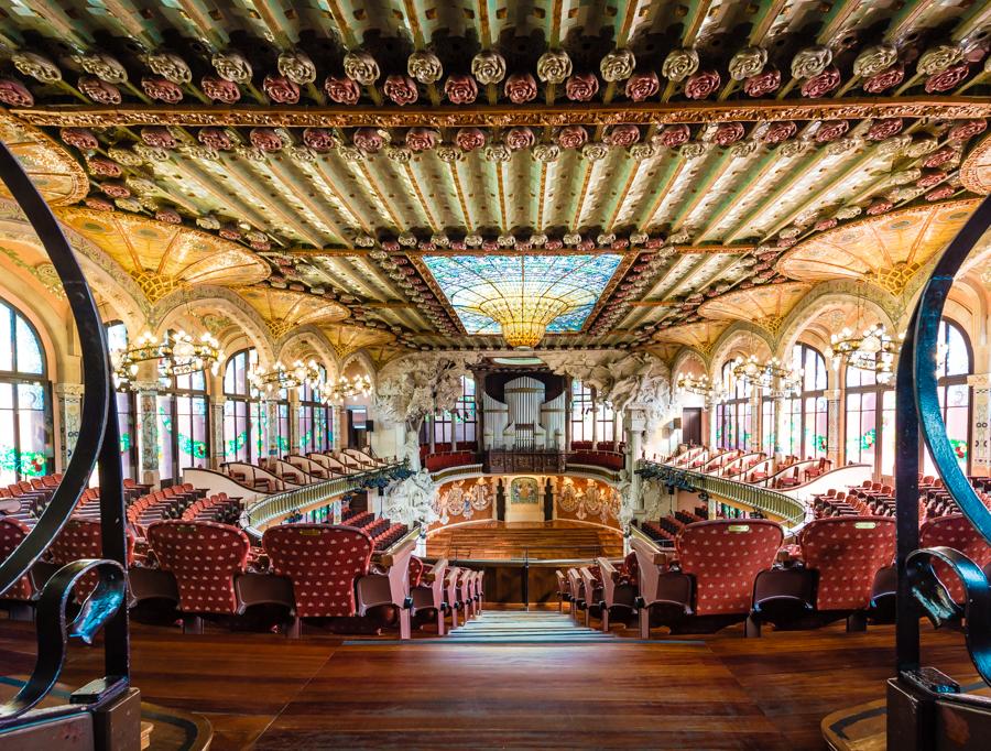 a large room with many seats and a stained glass ceiling with Palau de la Música Catalana in the background