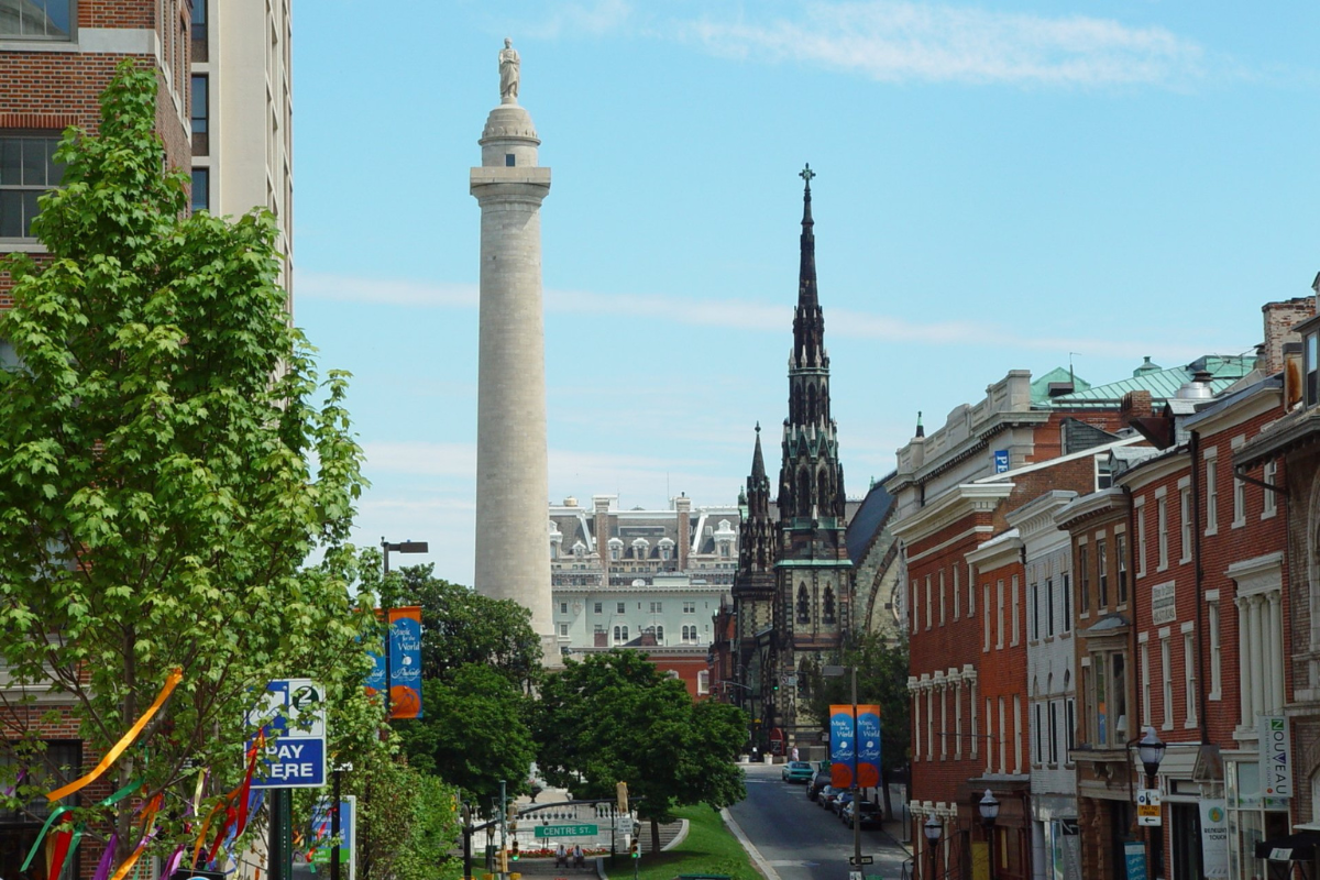 a tall white tower with a statue on top of it with Mount Vernon, Baltimore in the background