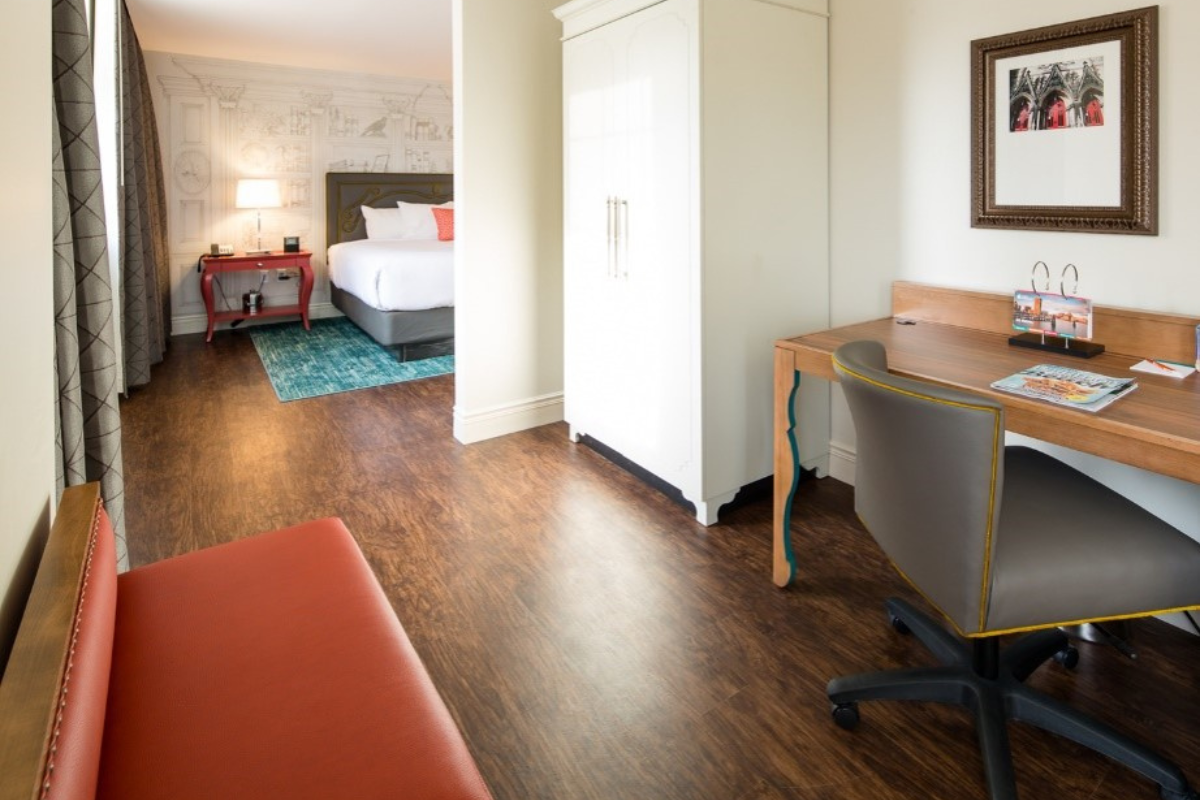 A modern hotel suite featuring dark wood laminate flooring, a study desk with an office chair, a white wardrobe, a red bench in the foreground, and an adjoining bedroom with a white bed and teal area rug.