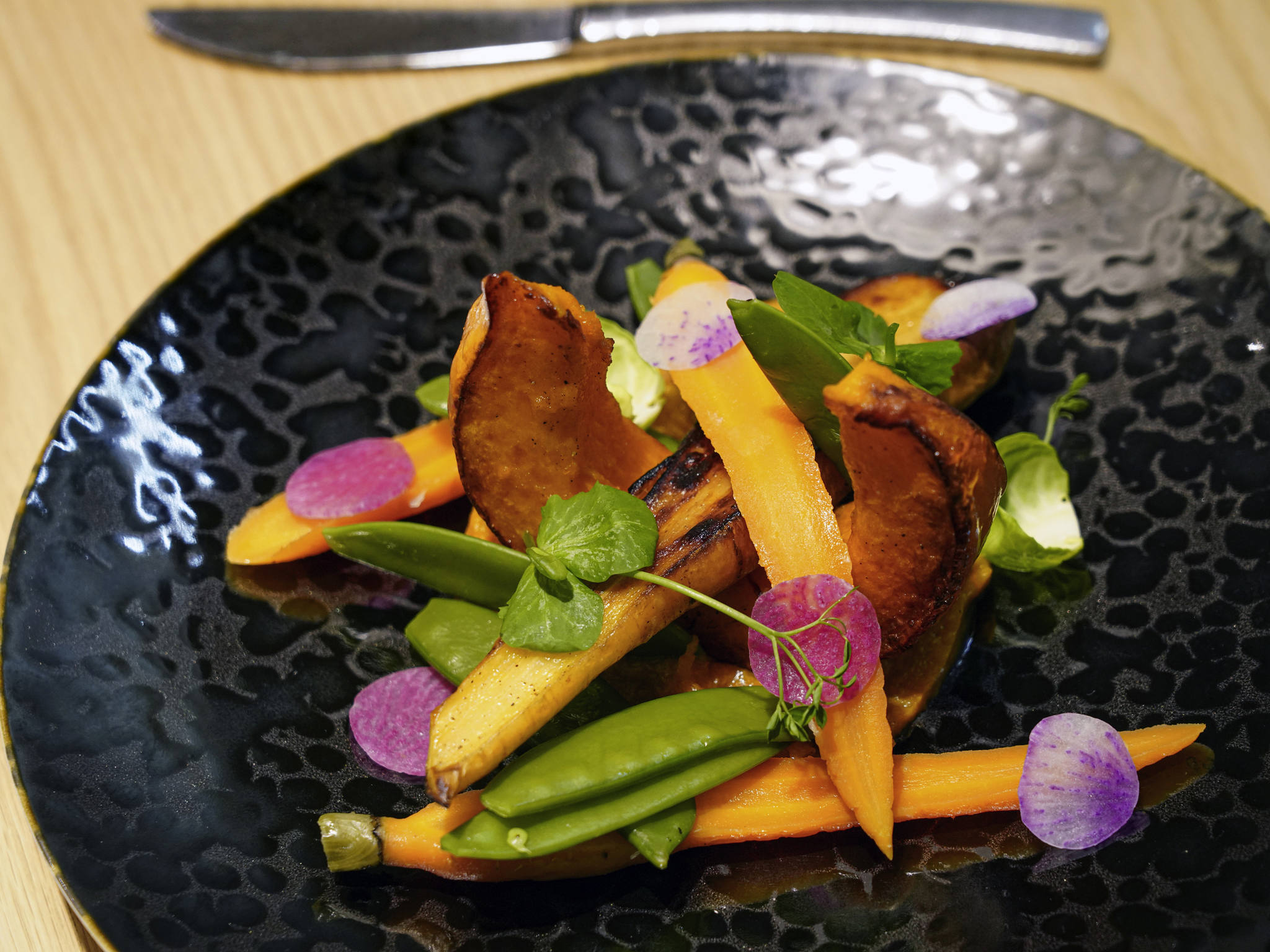 Artfully plated gourmet dish featuring roasted squash wedges, whole roasted baby carrots, snap peas, fresh microgreens, and purple radish slices arranged on a textured black ceramic plate.