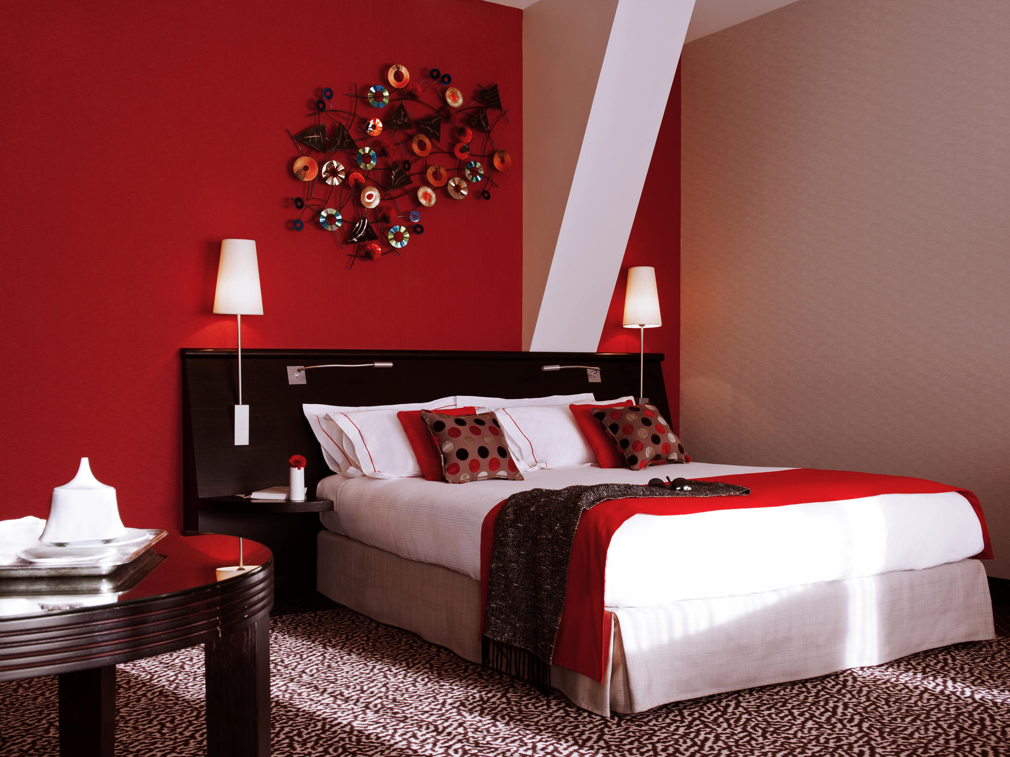 Modern hotel bedroom with a bold red feature wall, dark wooden headboard with wall-mounted lamps, crisp white bedding accented with red throw pillows and runner, a patterned carpet, and colorful geometric metal wall art above the bed.