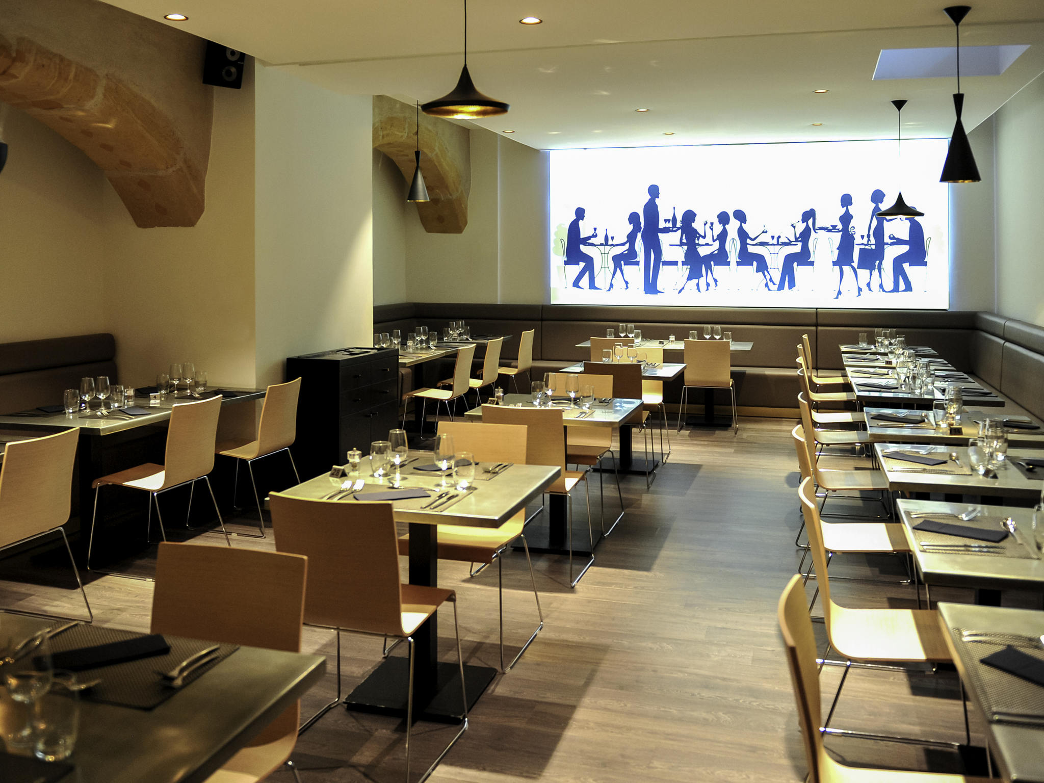 Modern hotel restaurant dining room with stone archways, light wood chairs, pendant lights, set tables, and a large illuminated mural depicting silhouettes of people dining.