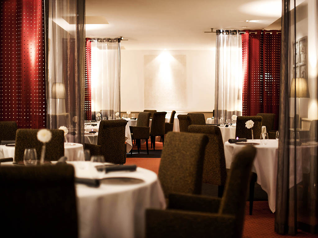 Upscale restaurant interior featuring round dining tables with white tablecloths, dark upholstered chairs, warm ambient lighting, and sheer dark and red curtains dividing the seating areas.