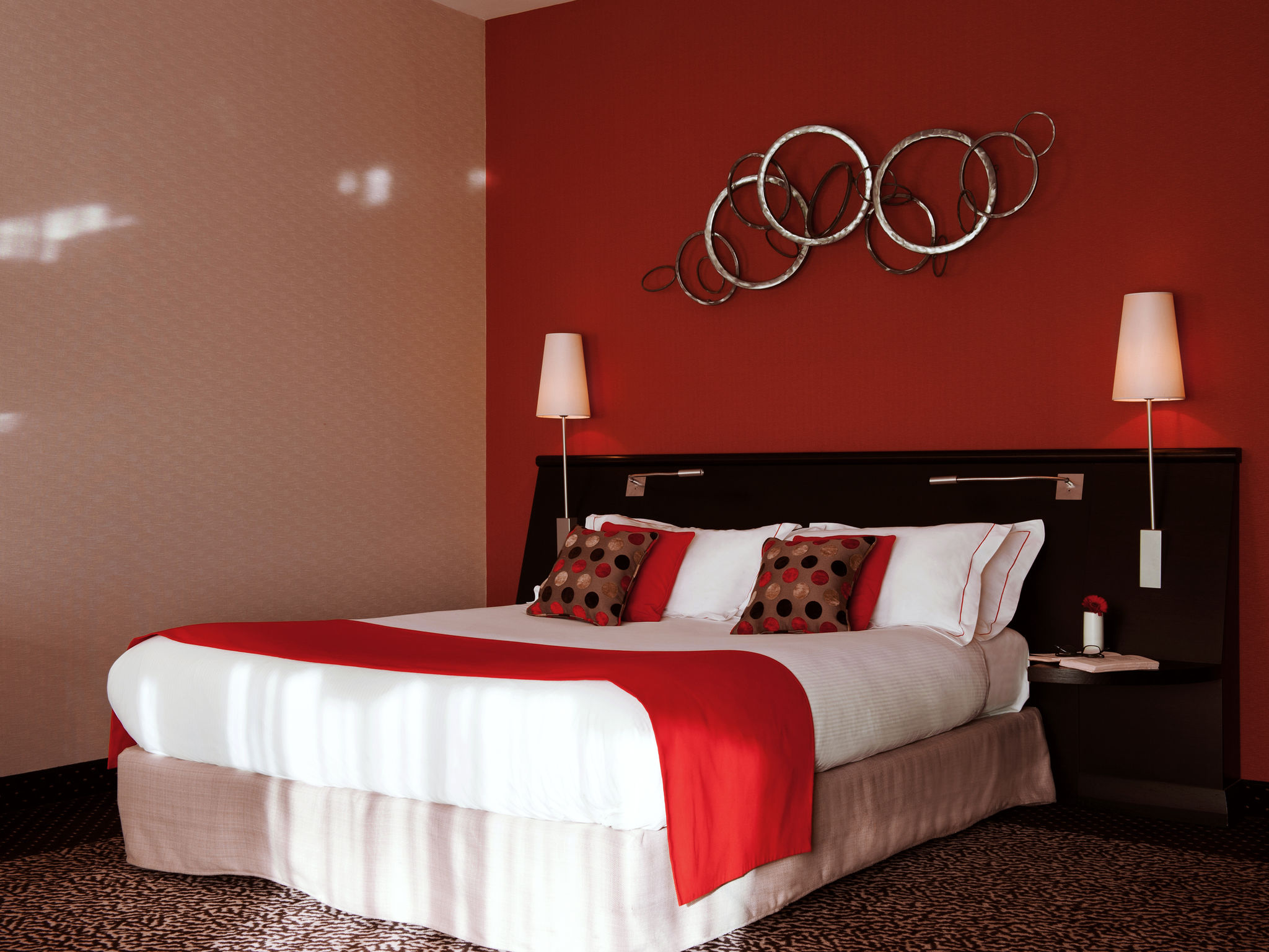 Contemporary hotel guest room featuring a plush king bed with crisp white bedding, a bright red accent runner, matching red throw pillows, a dark wood headboard with integrated bedside lights, a bold red accent wall adorned with interlocking metal ring wa