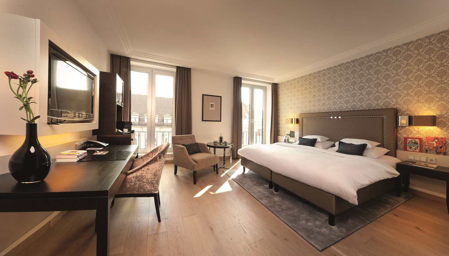 An elegant hotel room with warm wood flooring, a double bed set against a patterned feature wallpaper, a dark wood work desk with a TV mounted above, an armchair near large French windows, and warm ambient wall lights.