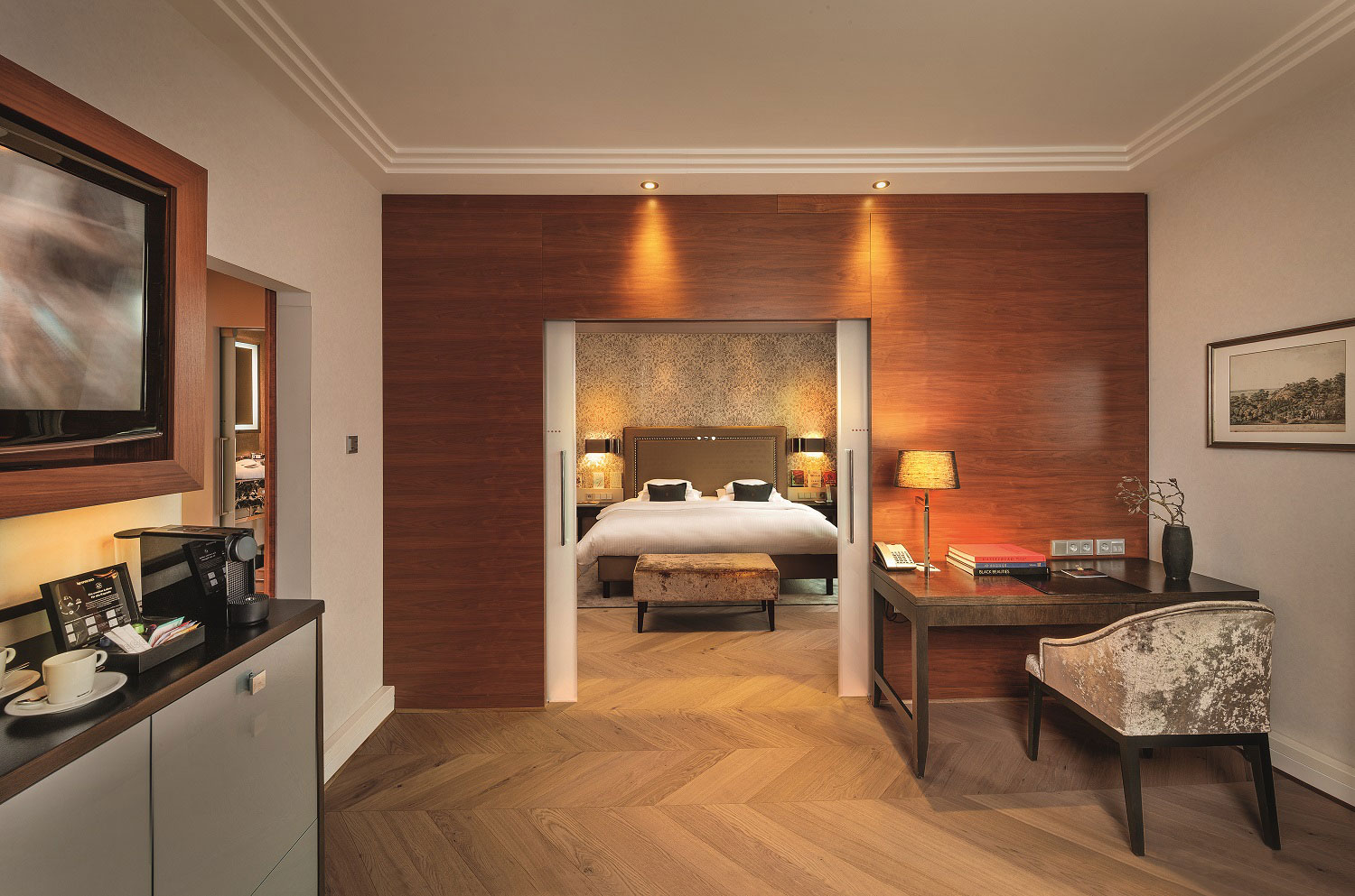 A modern hotel suite view looking through sliding glass doors past a warm wood feature wall into a cozy bedroom with chevron parquet floors, a writing desk with a velvet chair, and a coffee amenities station in the foreground.
