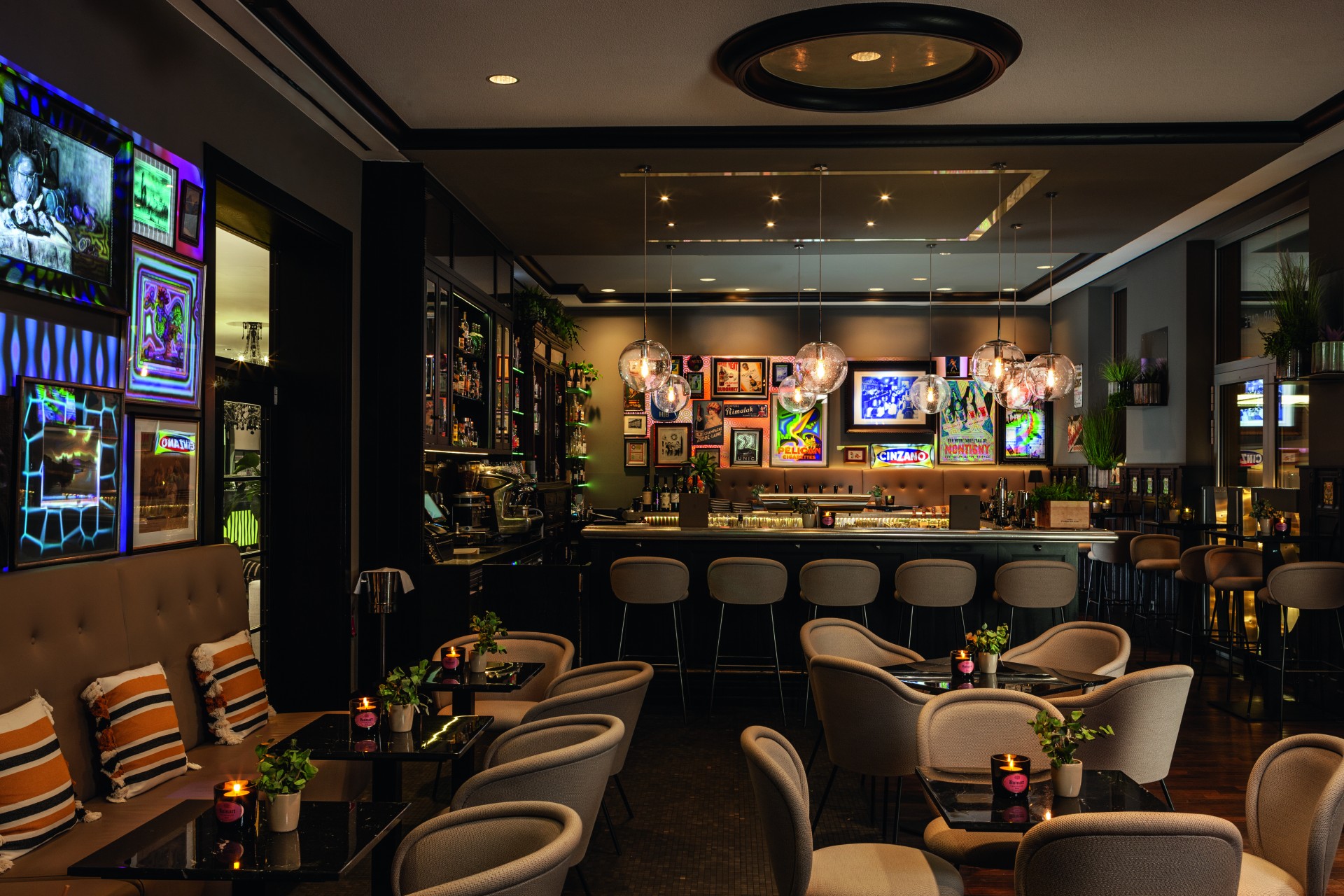 A chic, moody bar lounge featuring glowing pendant glass globes over the bar counter, beige upholstered tub chairs around small dark tables, and illuminated colorful artwork lining the walls.