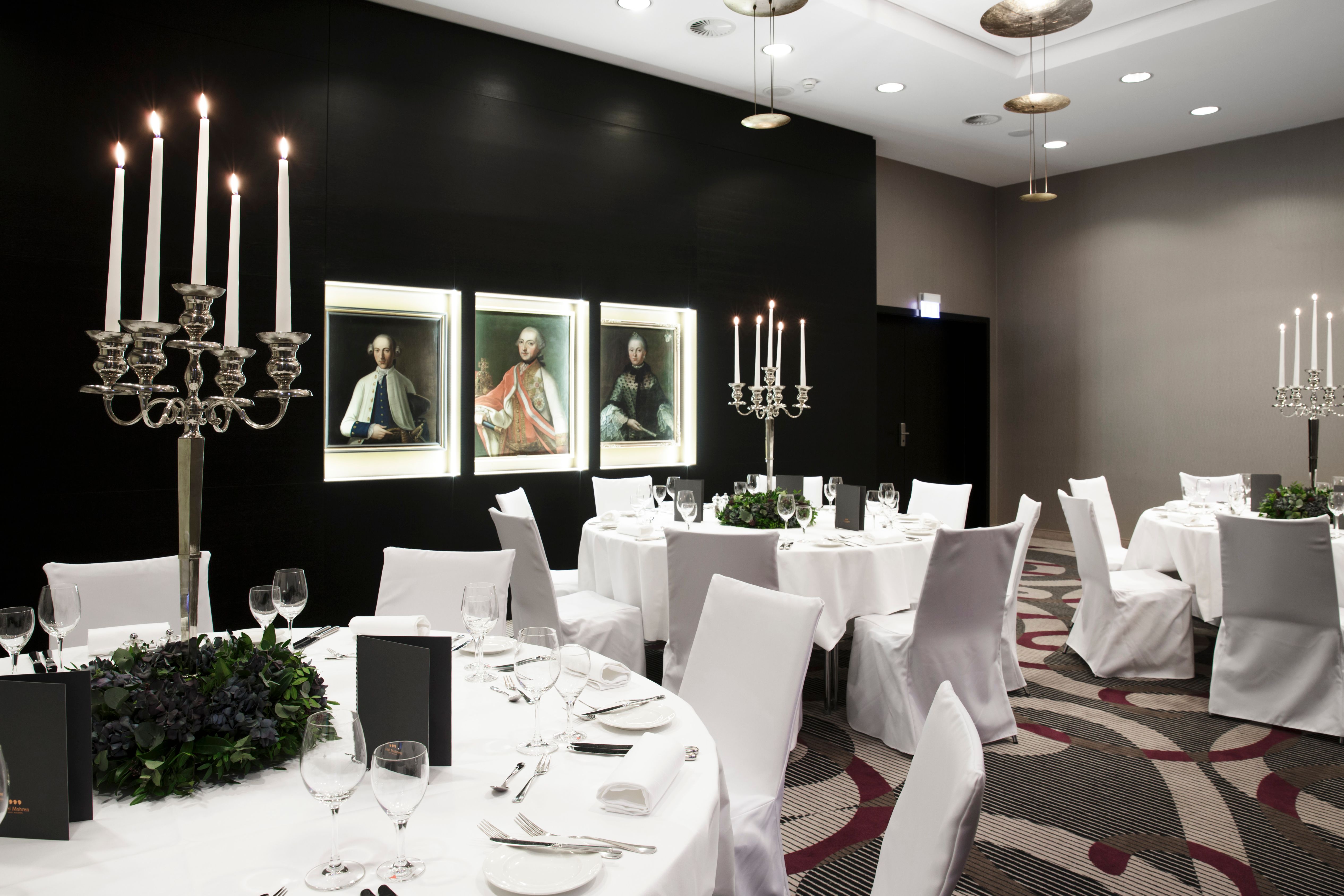 An elegant banquet hall setup with round tables dressed in white linens and chair covers, topped with floral centerpieces and tall silver candelabras, featuring illuminated classical portraits along a dark accent wall.