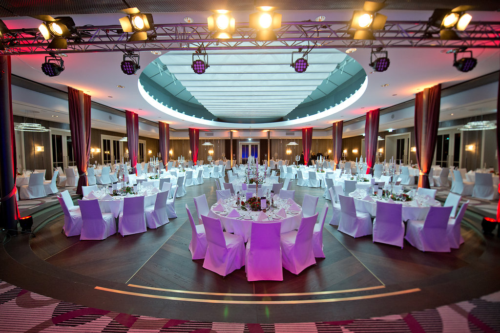 A vast, circular event space set up for a gala with round dining tables clothed in white, red ambient lighting accents, dramatic vertical drapery, and overhead stage lighting rig below a skylight.