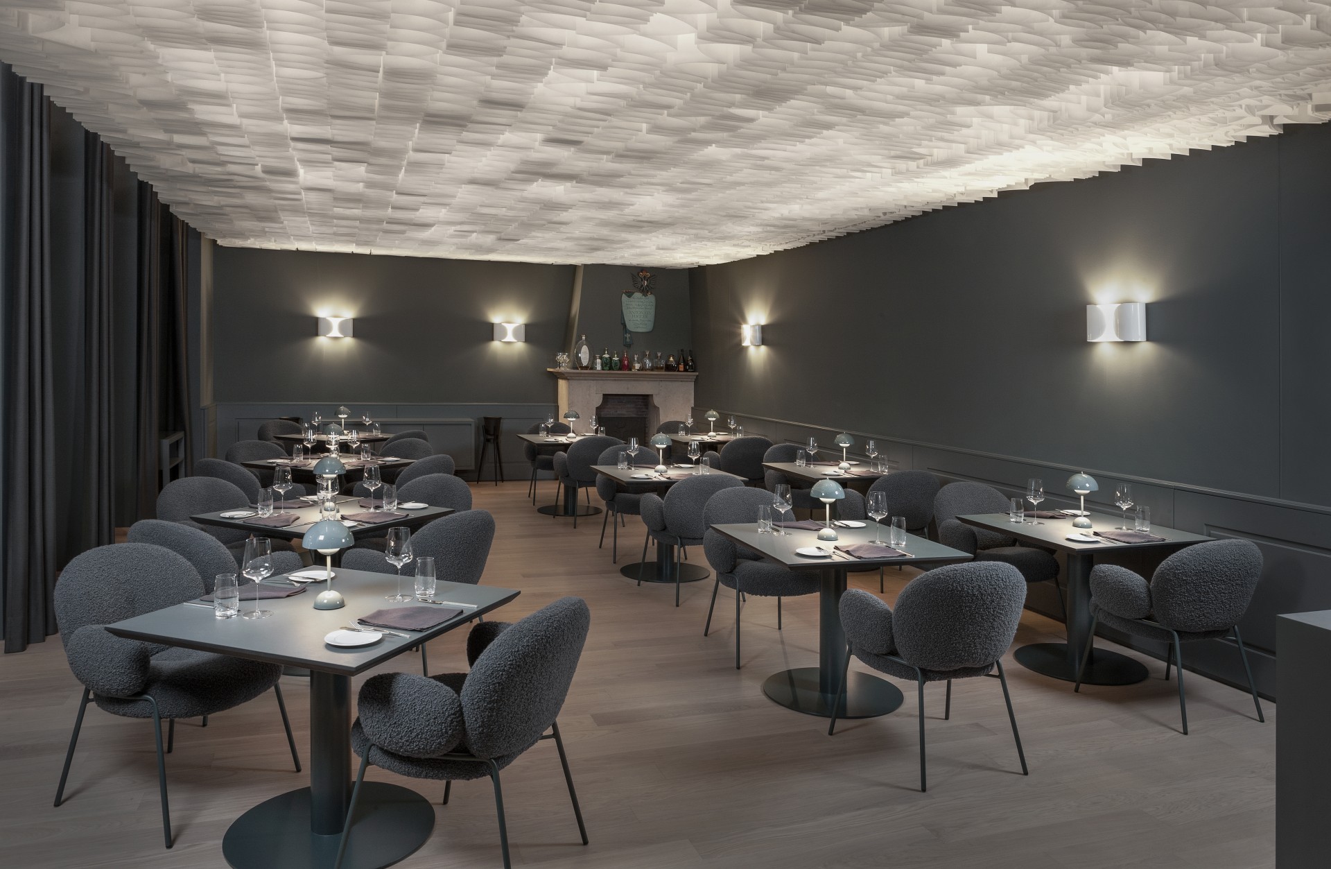 A sleek, modern restaurant dining room featuring dark grey walls, dark textured armchairs, small pedestal tables with miniature lamps, and a striking illuminated textured ceiling installation.