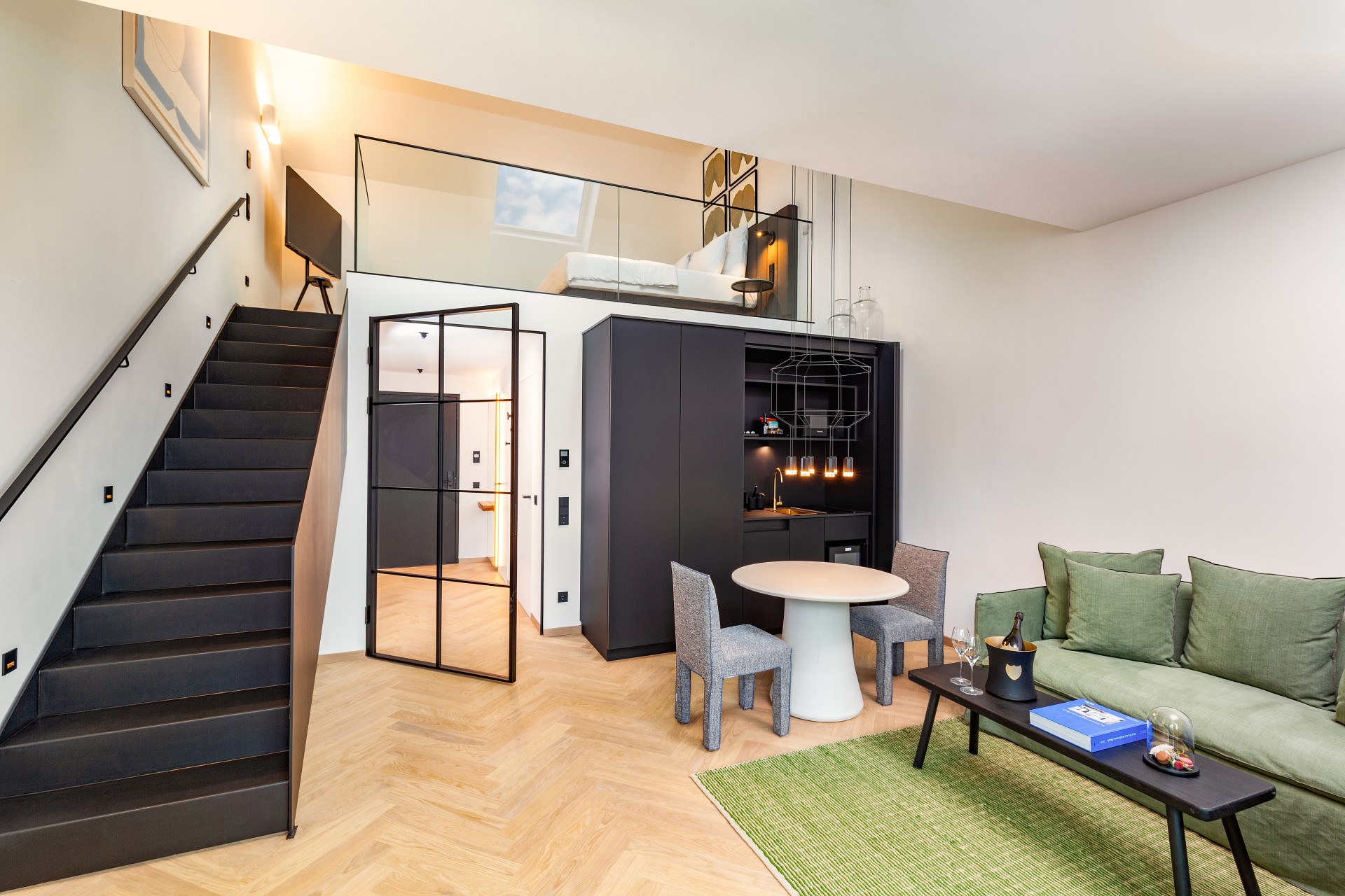 A contemporary two-story loft suite with light wood herringbone floors, featuring a lower-level living area with a sage green sofa, small dining set, and dark kitchenette, leading up black stairs to a mezzanine bedroom with glass railings.