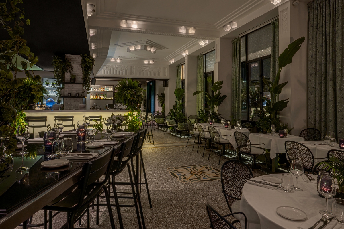 A stylish, dimly lit restaurant interior featuring high-top bar seating on the left, white-clothed tables with black mesh chairs on the right, terrazzo flooring with a central tile motif, and lush indoor greenery throughout.