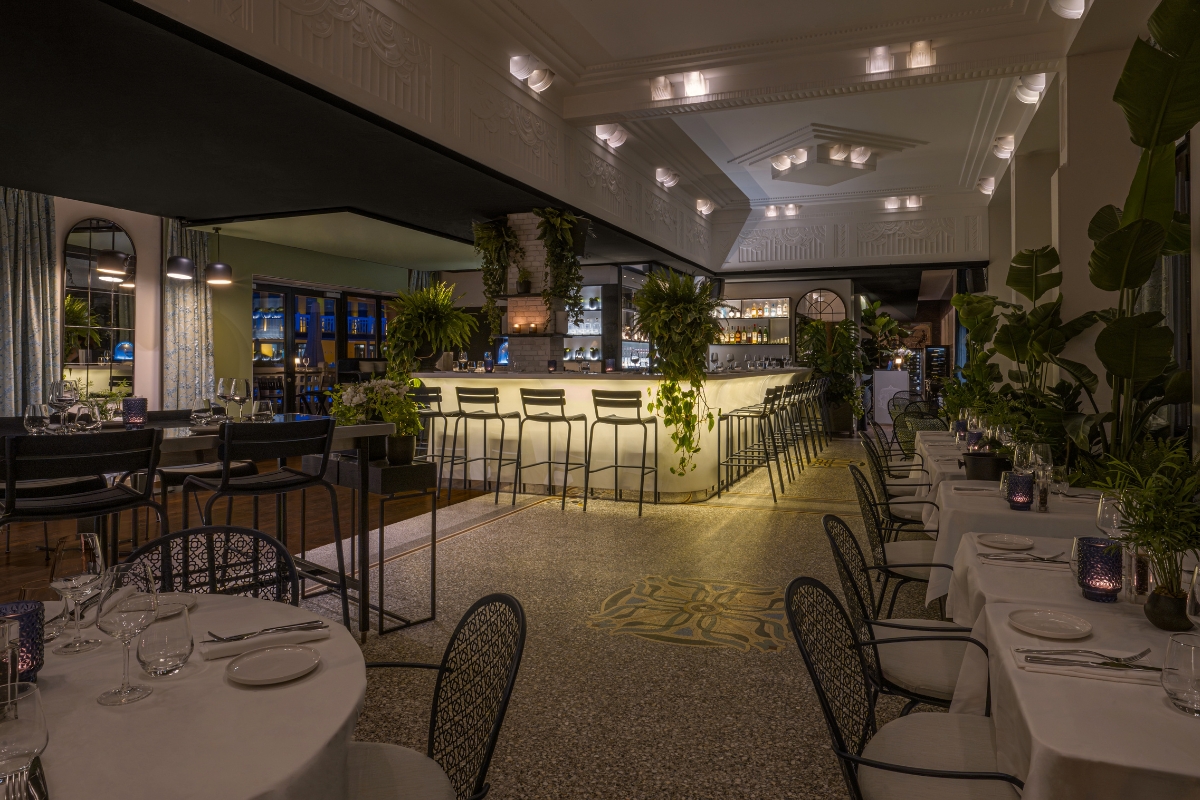A wider view of a sophisticated, mood-lit hotel restaurant and bar with a brightly illuminated bar counter, white-clothed dining tables, dark wire mesh armchairs, terrazzo floors, and leafy tropical potted plants.