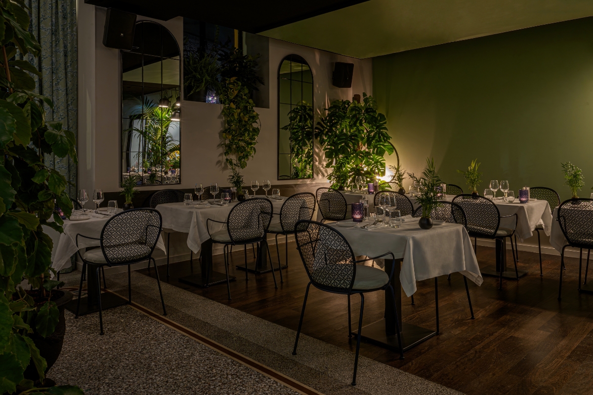 An intimate restaurant dining area with dark wooden floors, olive green walls, small tables set with white tablecloths, patterned mesh chairs, arched mirrors, and lush potted plants creating a cozy atmosphere.