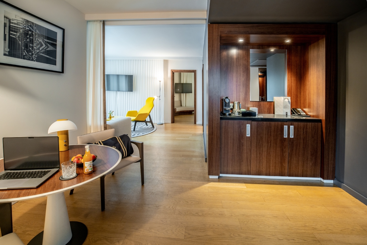 A modern, spacious hotel suite featuring light wood floors, a small desk area with a laptop and fruit, a built-in dark wood minibar, and a bright living area with a bold yellow armchair visible in the background.
