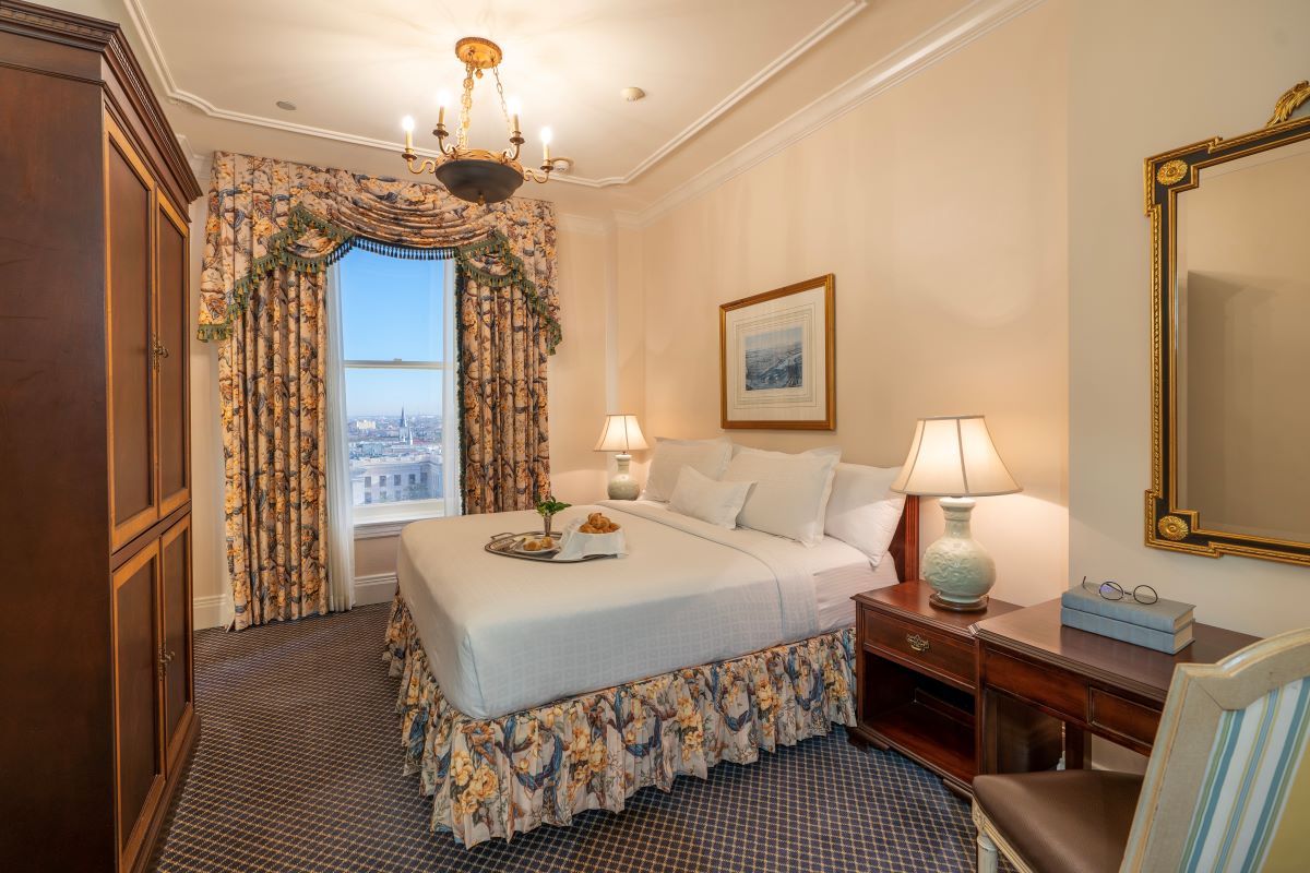 Classic hotel bedroom with a king bed dressed in white linens and a floral bed skirt, matching patterned drapes framing a window with city views, dark wood armoire, nightstands, and a hanging brass chandelier.