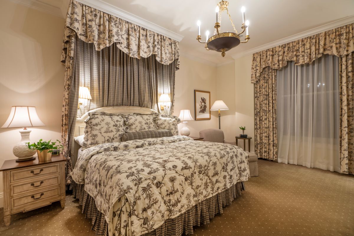 Traditional luxury hotel room featuring a bed with toile patterned bedding and matching draped canopy, bedside tables with lamps, a chandelier, and window drapes.