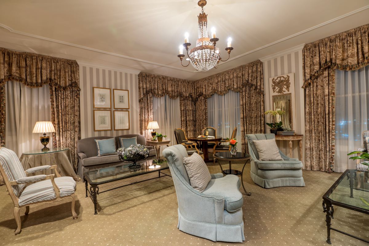Elegantly furnished luxury suite sitting room featuring classical drapes, a crystal chandelier, striped wallpaper, plush armchairs, sofa, glass coffee table, and a small dining setup in the background.