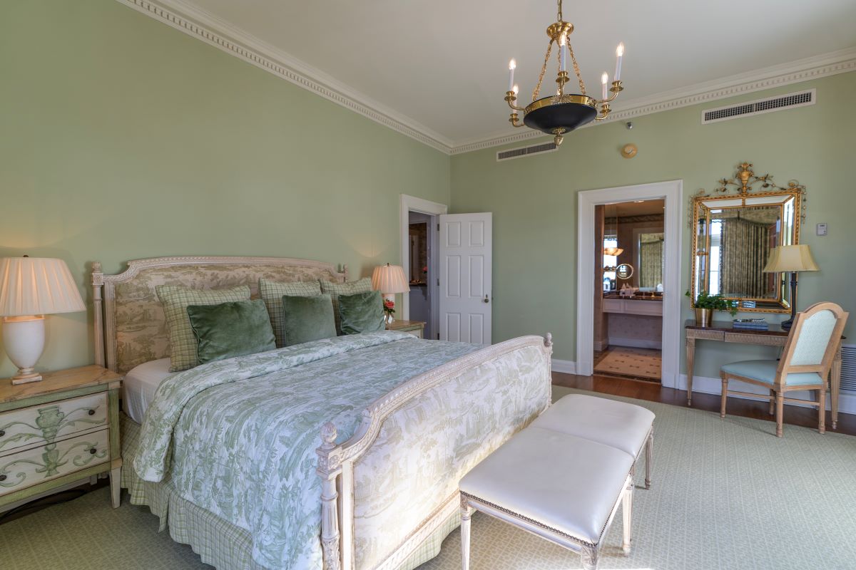 Elegant hotel bedroom featuring sage green walls, a upholstered bed with green patterned linens, nightstands with lamps, a chandelier, and a vanity set with an ornate mirror.