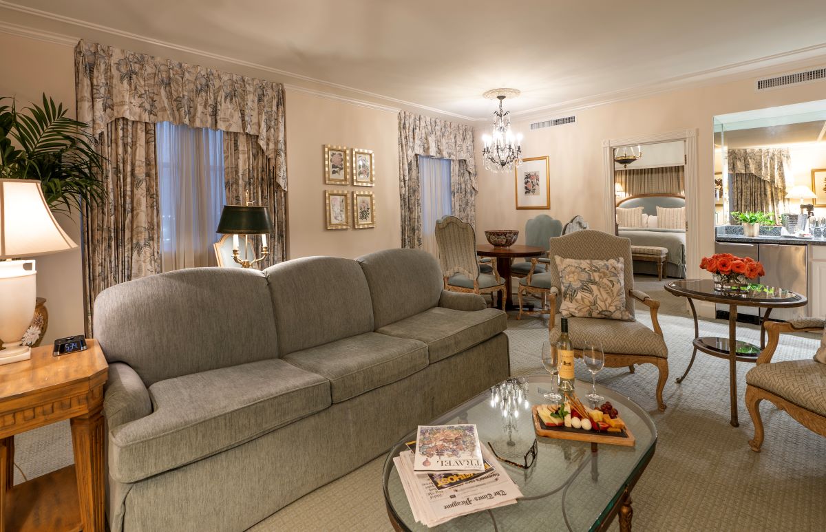 Spacious classic hotel suite living area with a long grey sofa, glass-top coffee table with wine and snacks, armchairs, floral drapes, and an adjacent bedroom visible through an open doorway.