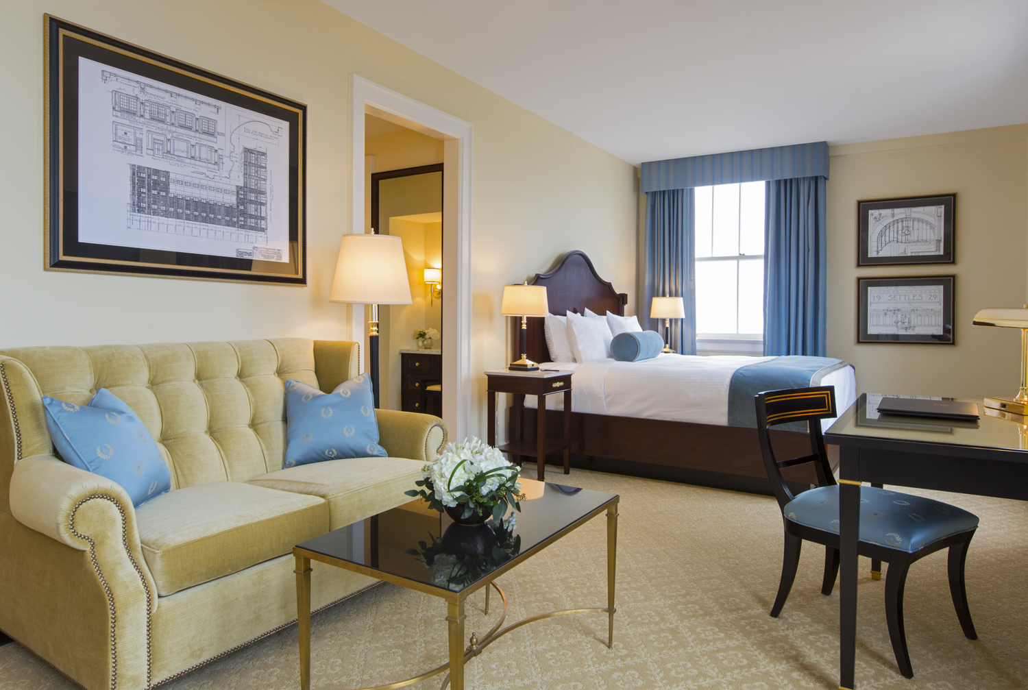 A hotel suite sitting area and bedroom featuring a tufted light-green sofa with nailhead trim, a black glass coffee table, framed architectural blueprint artwork, and a dark wood bed with white linens in the background near a window.