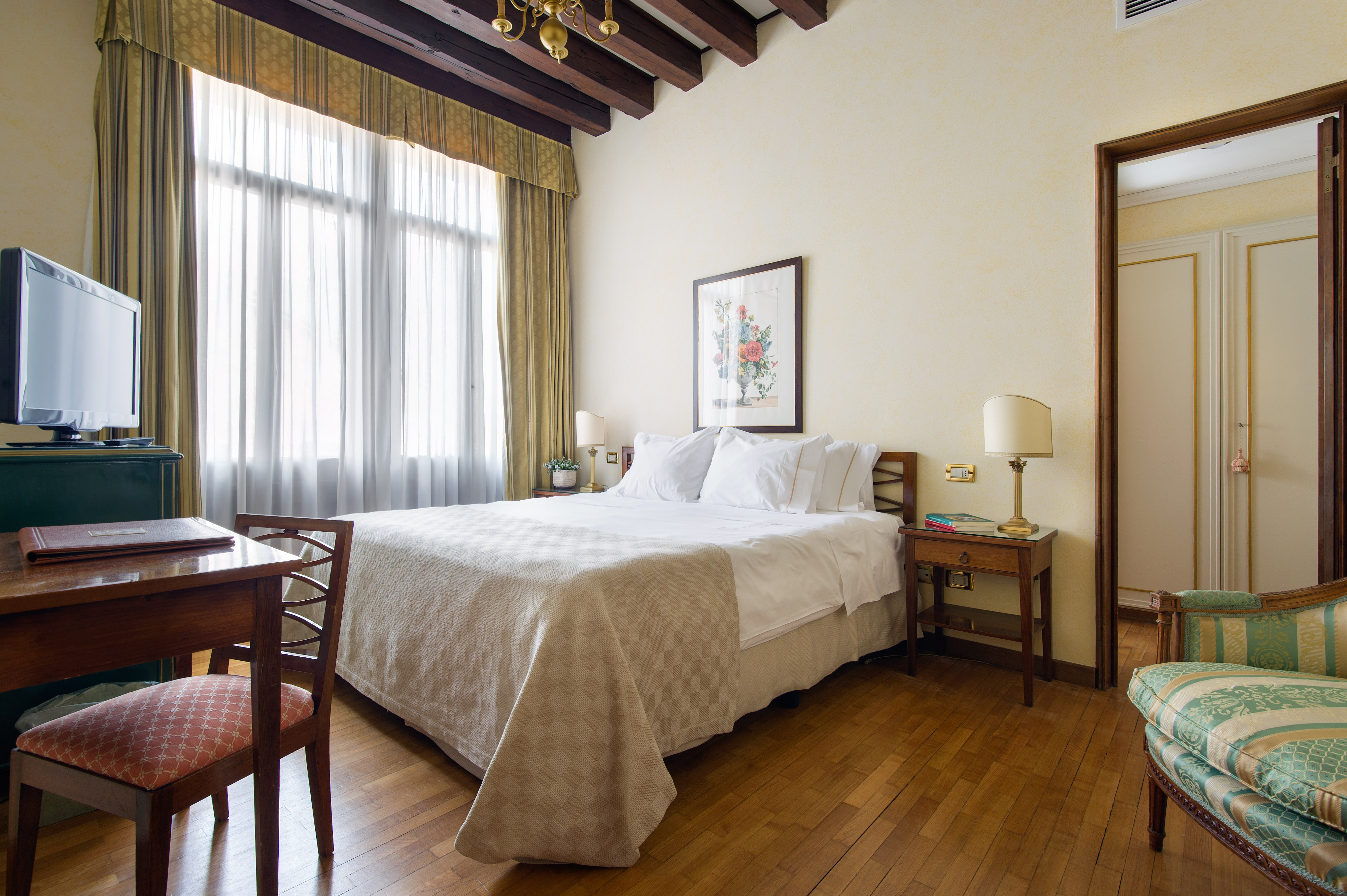A classic hotel bedroom with dark wood ceiling beams, a queen-sized bed dressed in white linens, a wooden desk with a chair, and a green patterned armchair in the foreground.