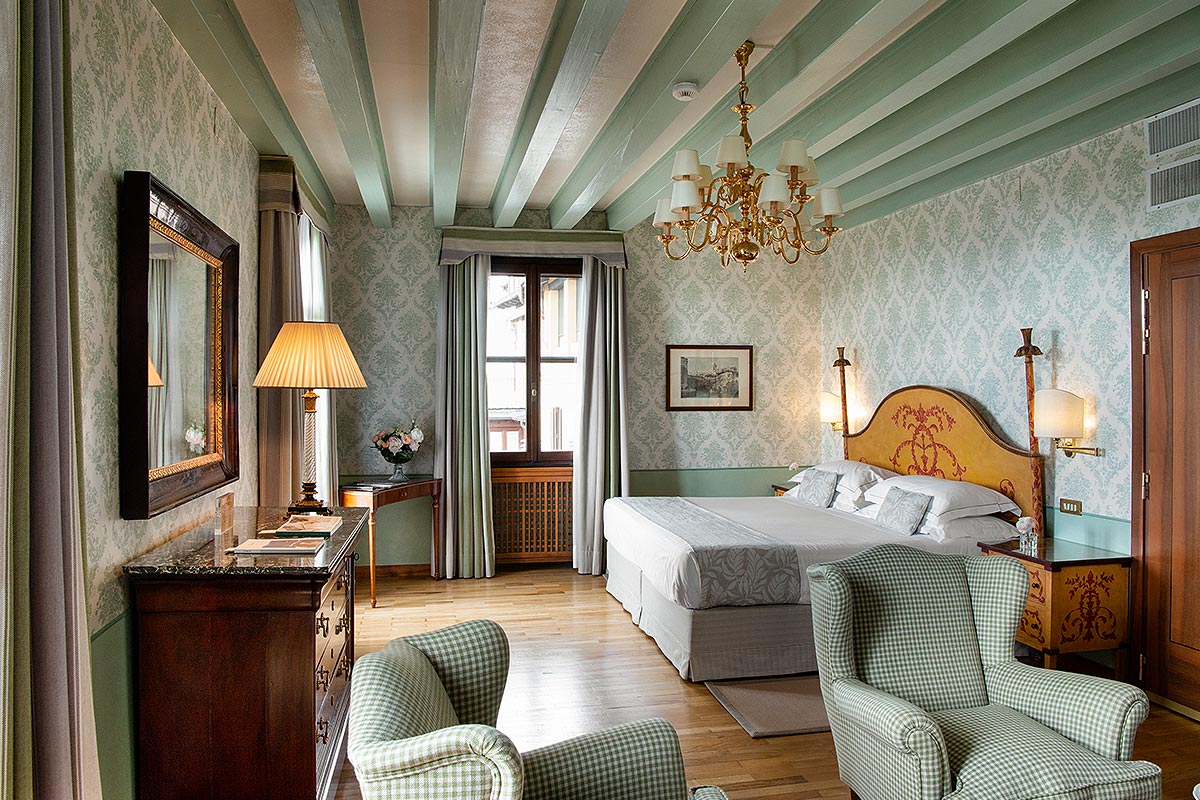 An elegant hotel room with sage green damask wallpaper, a matching beam ceiling, a large bed with a yellow ornate headboard, brass chandelier, and green gingham armchairs.