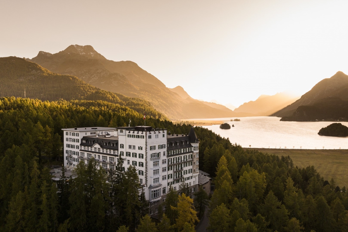 An aerial view of a large, grand white hotel building nestled among dense evergreen forests, set against a stunning mountain backdrop and a picturesque lake bathed in warm sunset light.