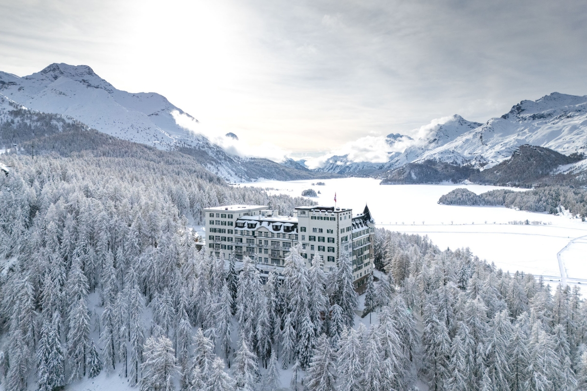 An aerial view of a grand white historic hotel nestled within a snow-covered evergreen forest, overlooking a frozen lake and snow-capped alpine mountains under a bright sky.
