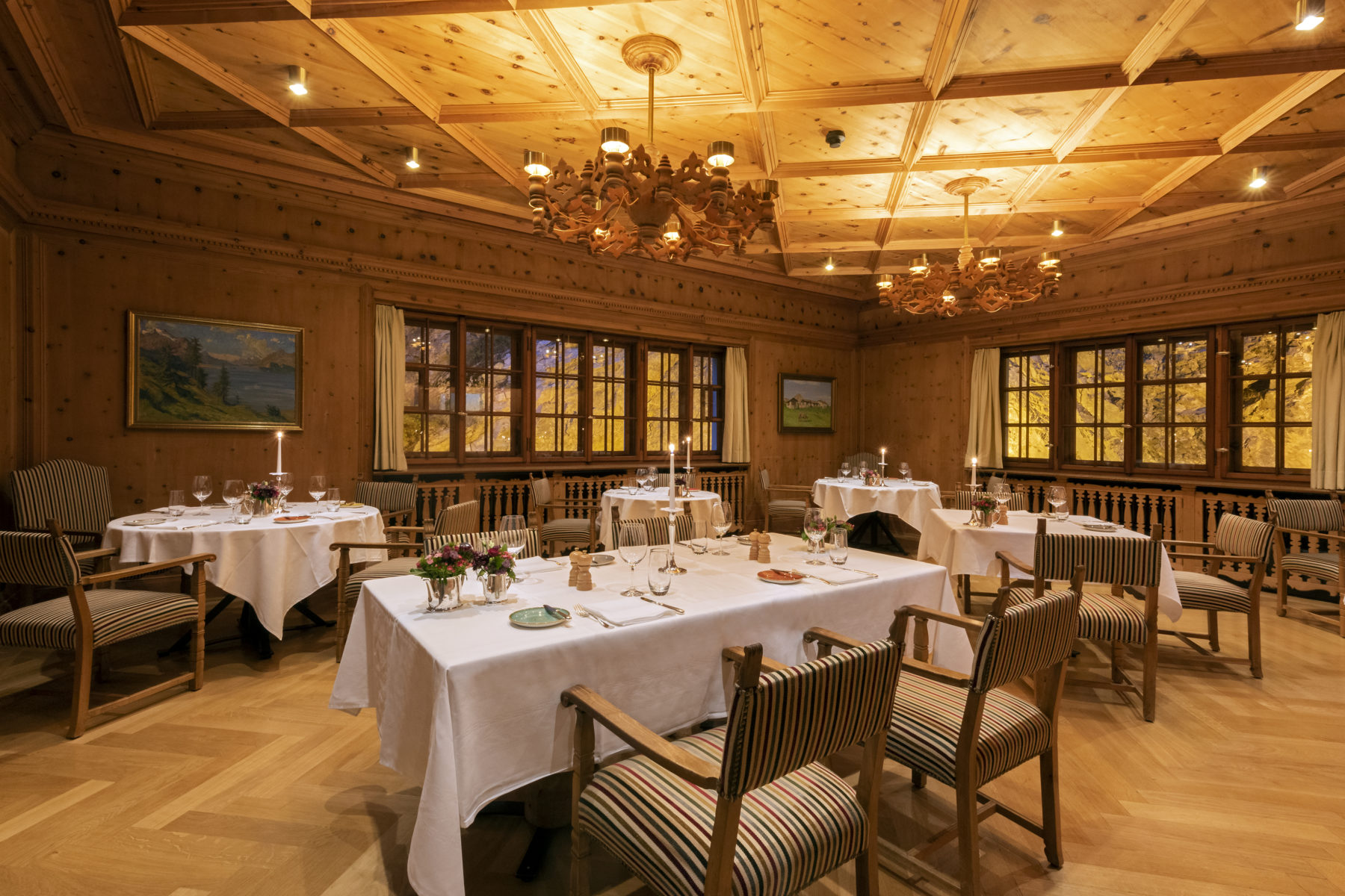 A cozy, alpine-style fine dining room with natural wood-paneled walls and an intricately coffered wooden ceiling. Tables dressed in crisp white tablecloths are set with wine glasses, lit candles, and floral arrangements, surrounded by striped upholstered 